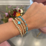 A close-up of a person's wrist wearing three beaded stretch bracelets: two gold-toned with white beads and one turquoise blue with a small cross charm.