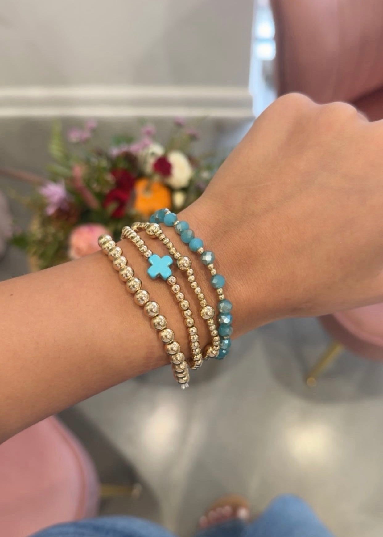 A close-up of a person's wrist wearing three beaded stretch bracelets: two gold-toned with white beads and one turquoise blue with a small cross charm.