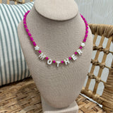A beaded necklace with bright pink beads and white star and letter charms spelling 'MOTHER', displayed on a beige fabric jewelry bust in a wicker chair setting.