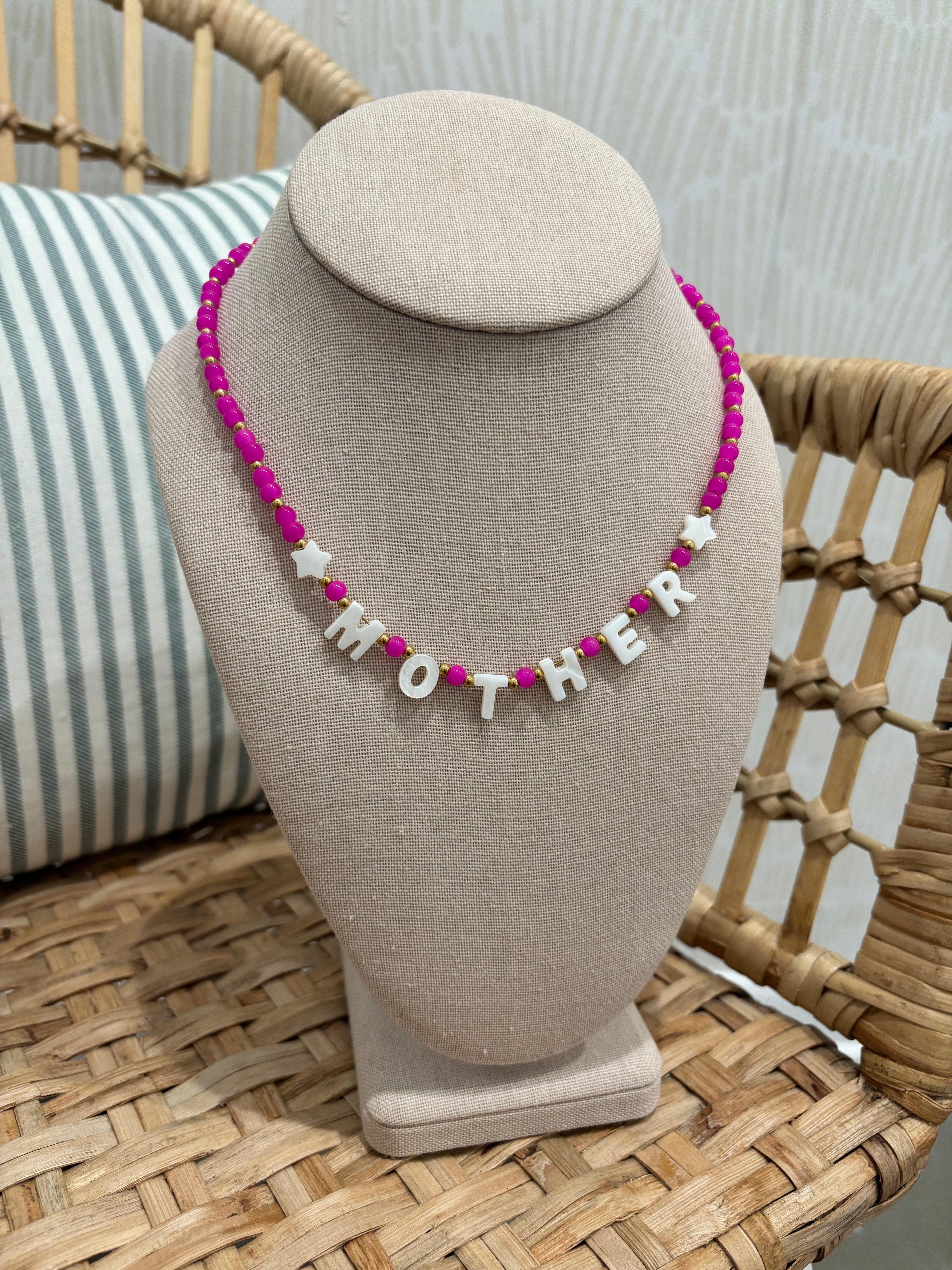 A beaded necklace with bright pink beads and white star and letter charms spelling 'MOTHER', displayed on a beige fabric jewelry bust in a wicker chair setting.