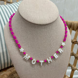 A beaded necklace displayed on a beige fabric jewelry bust. The necklace features bright pink round beads, small gold spacer beads, and white letter beads spelling 'MOTHER' with white star beads.