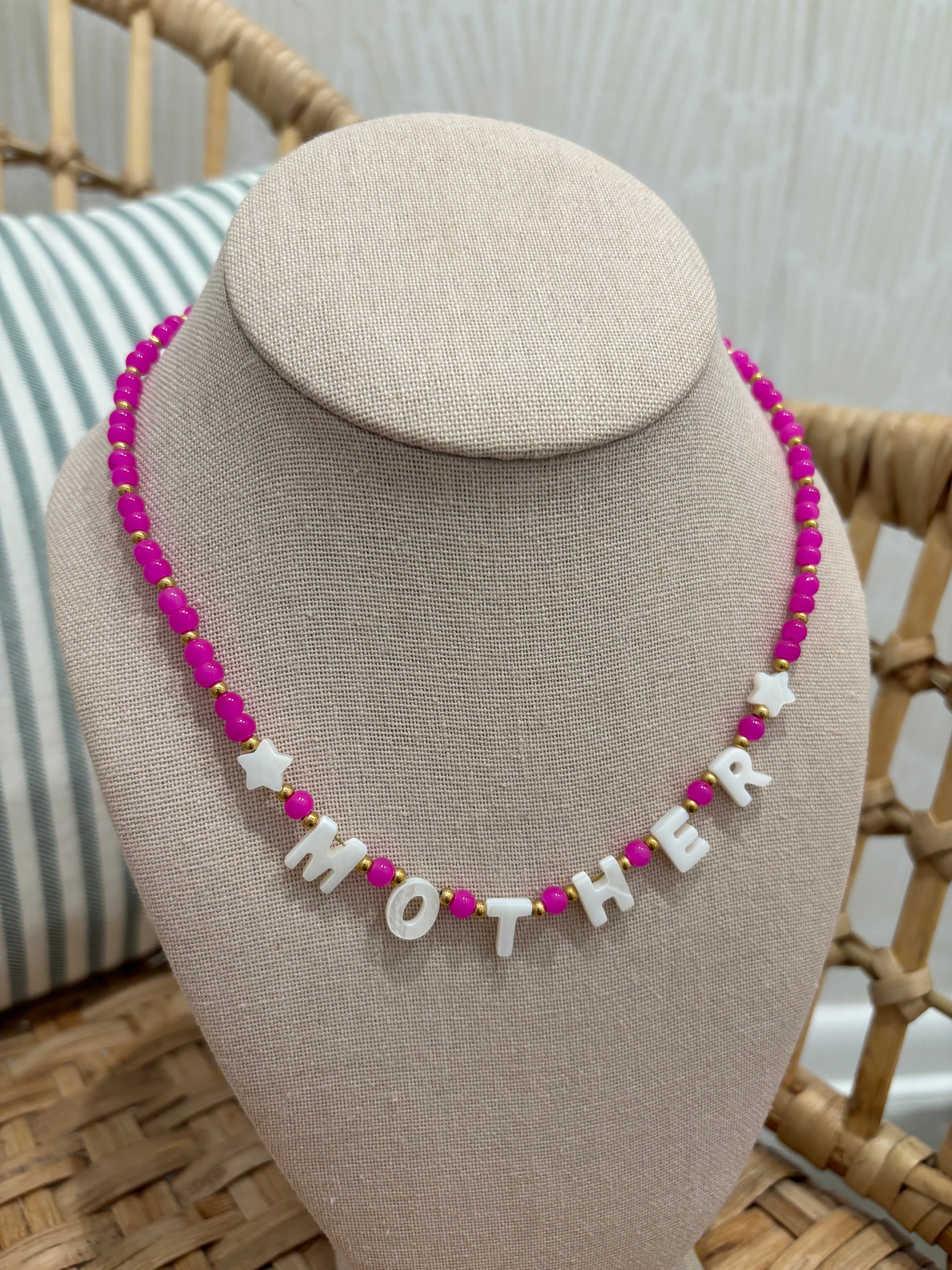 A beaded necklace displayed on a beige fabric jewelry bust. The necklace features bright pink round beads, small gold spacer beads, and white letter beads spelling 'MOTHER' with white star beads.