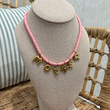 A pink cord necklace with gold letter charms spelling 'MOTHER' displayed on a beige fabric jewelry bust, set on a woven rattan chair with a striped pillow in the background.