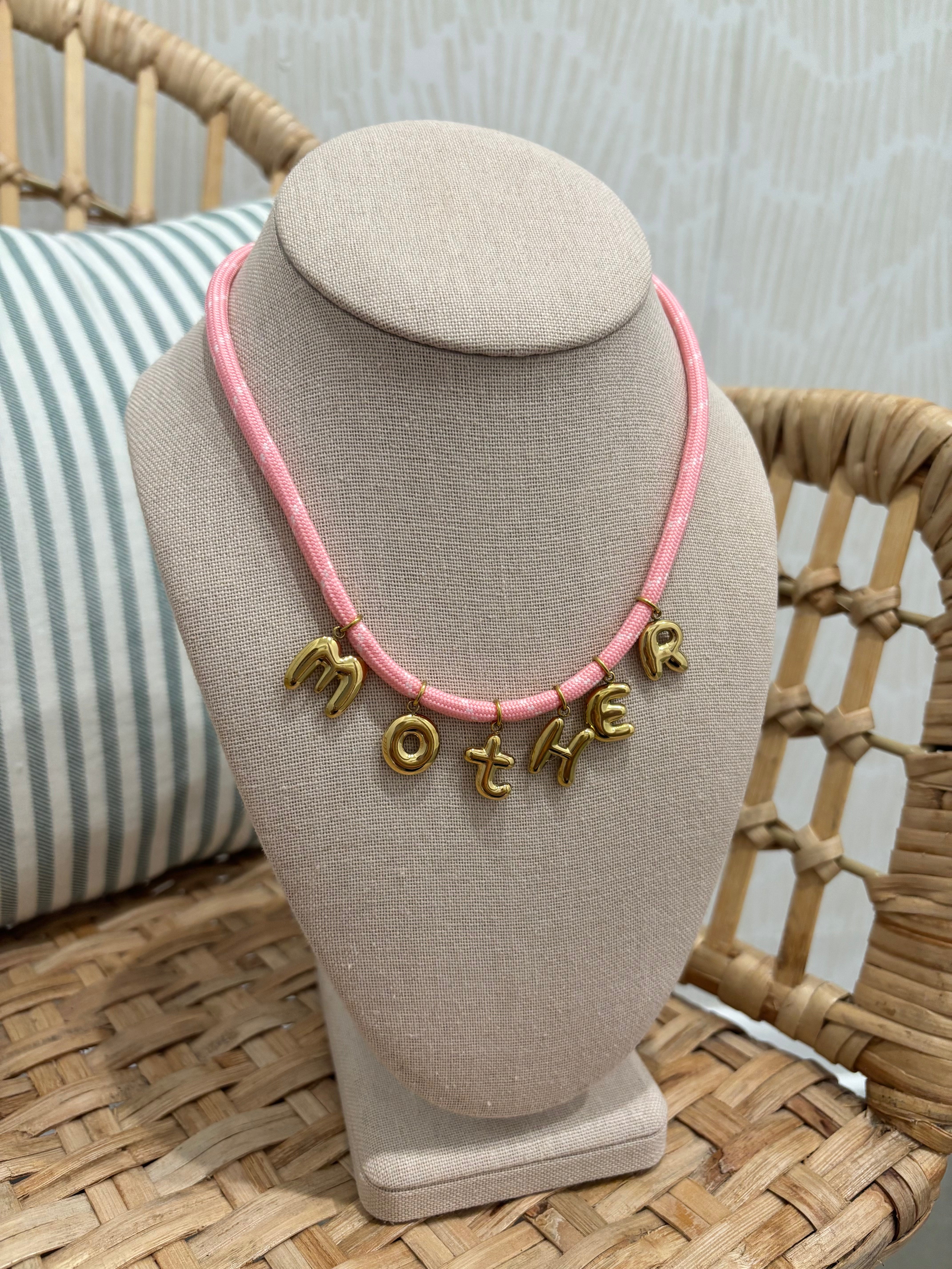 A pink cord necklace with gold letter charms spelling 'MOTHER' displayed on a beige fabric jewelry bust, set on a woven rattan chair with a striped pillow in the background.