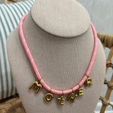 A pink braided cord necklace with gold letter charms spelling 'MOTHER', displayed on a beige fabric jewelry bust.