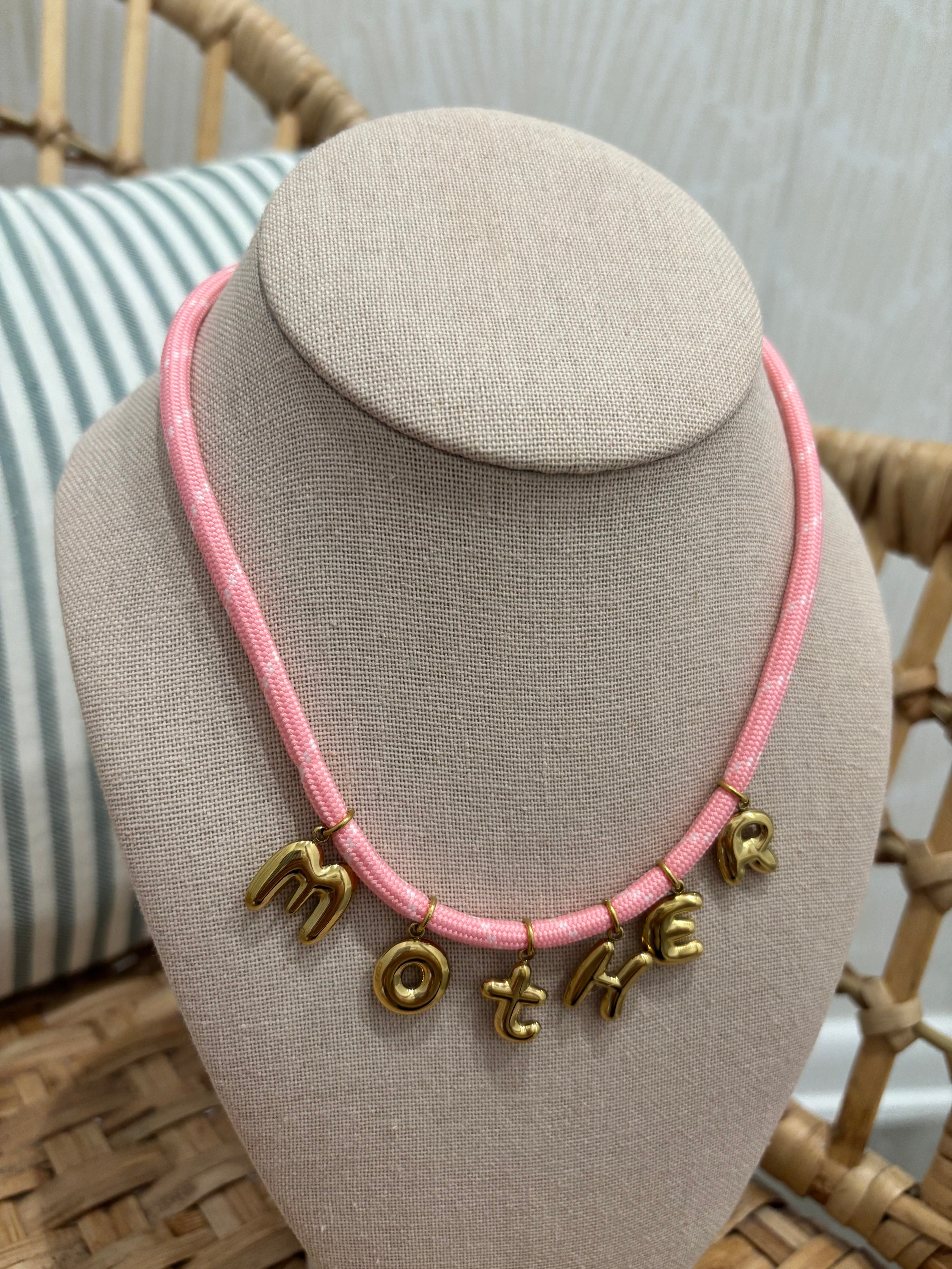 A pink braided cord necklace with gold letter charms spelling 'MOTHER', displayed on a beige fabric jewelry bust.
