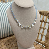 A personalized beaded necklace spelling 'MOTHER' with star accents, displayed on a beige fabric jewelry bust. The necklace features light blue faceted beads and white letter beads, set against a background of a woven rattan chair and a striped pillow.