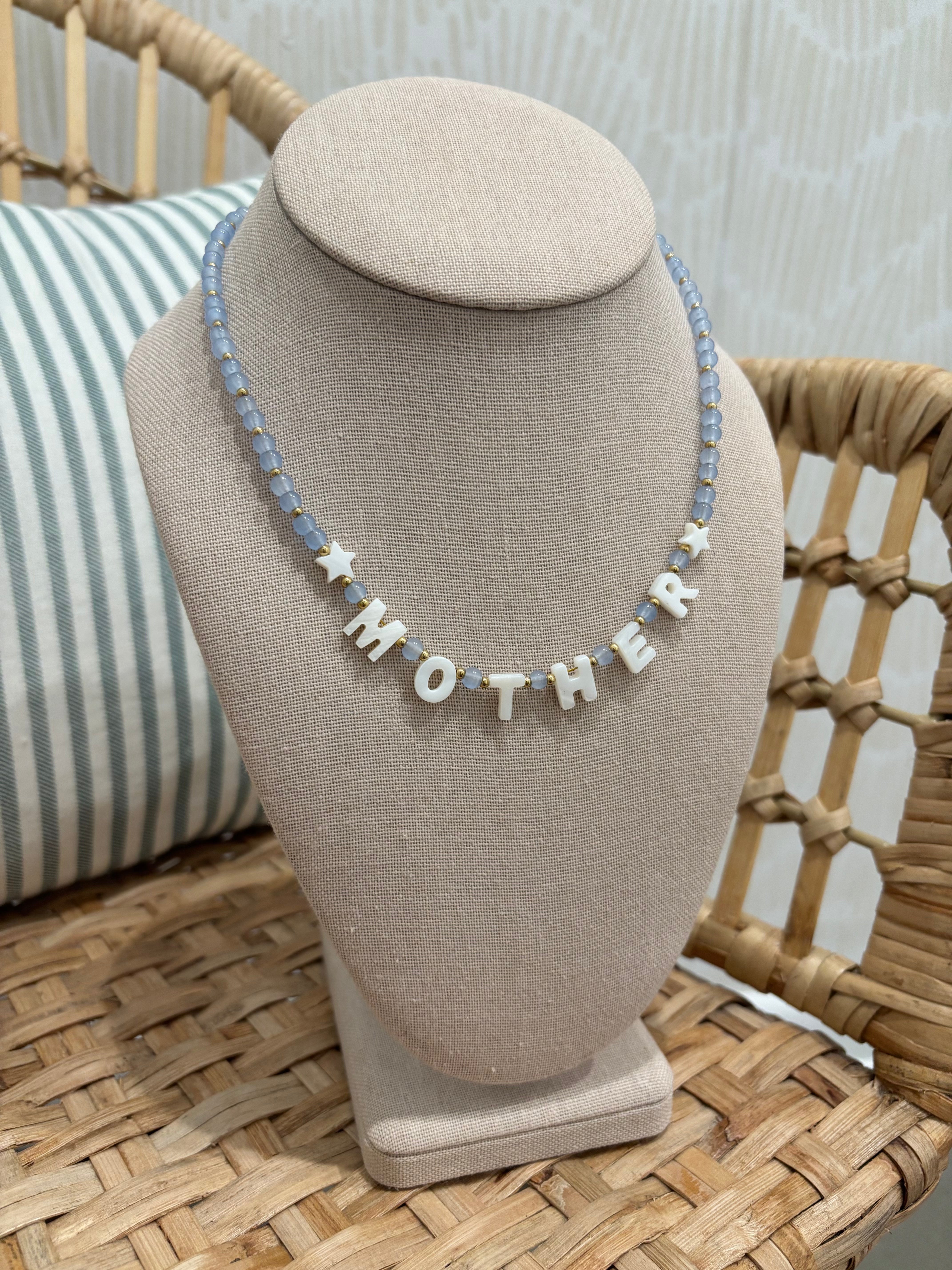 A personalized beaded necklace spelling 'MOTHER' with star accents, displayed on a beige fabric jewelry bust. The necklace features light blue faceted beads and white letter beads, set against a background of a woven rattan chair and a striped pillow.