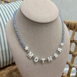 A beaded necklace displayed on a beige fabric jewelry bust. The necklace features light blue faceted beads, small white star-shaped beads, and white letter beads spelling 'MOTHER'.
