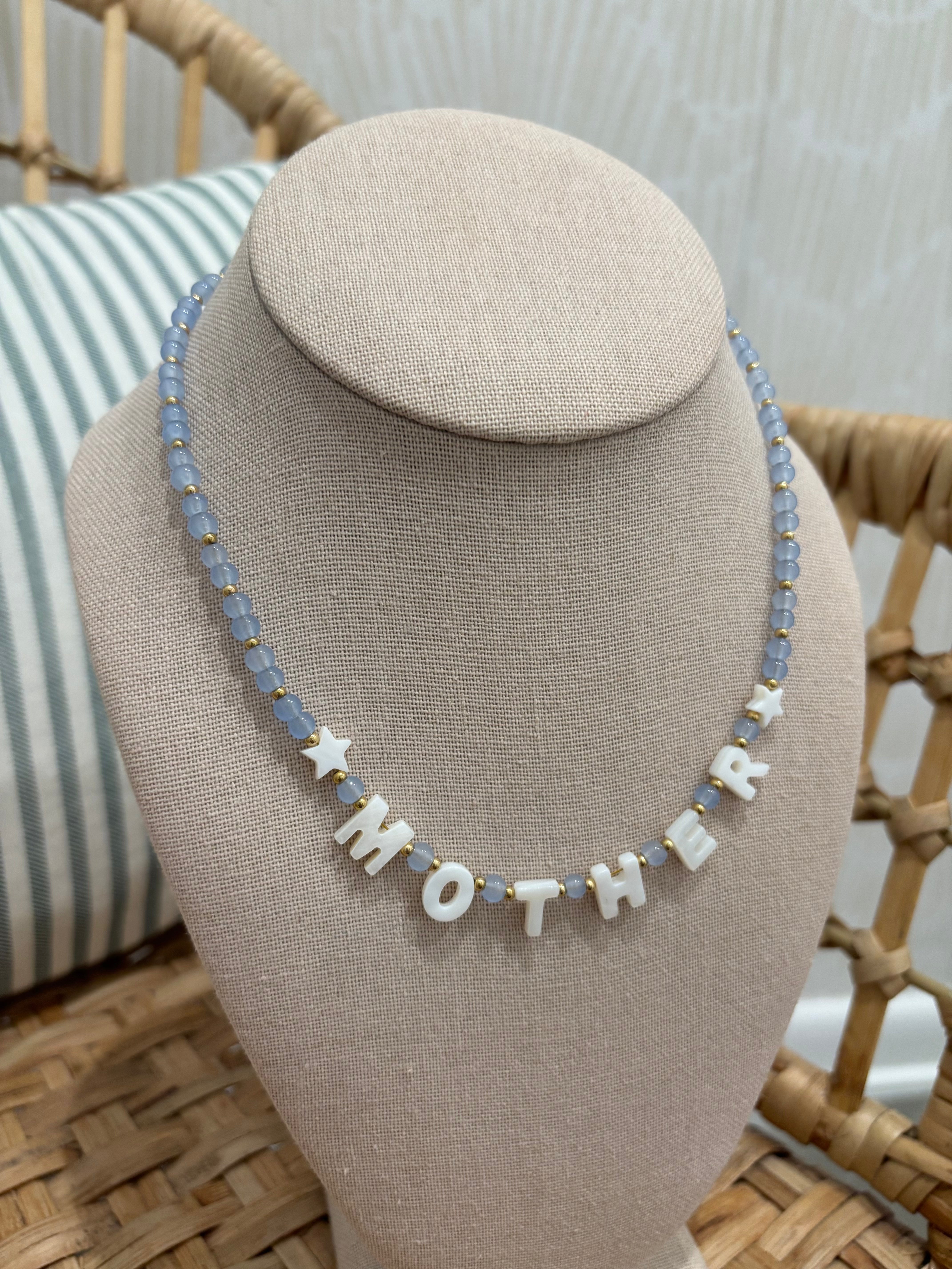A beaded necklace displayed on a beige fabric jewelry bust. The necklace features light blue faceted beads, small white star-shaped beads, and white letter beads spelling 'MOTHER'.