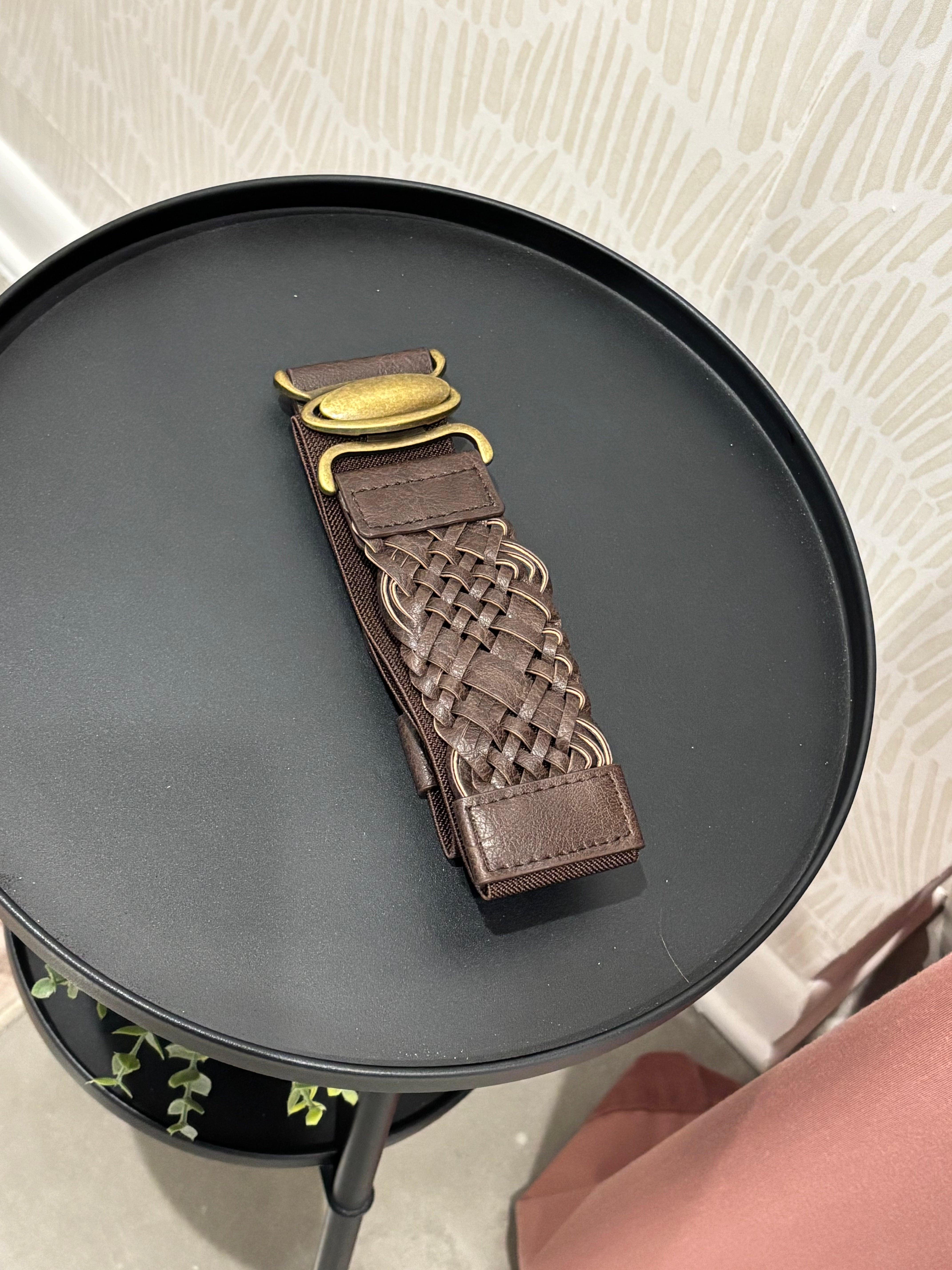A brown leather belt with a braided woven center and a gold-tone buckle, displayed on a round black side table.