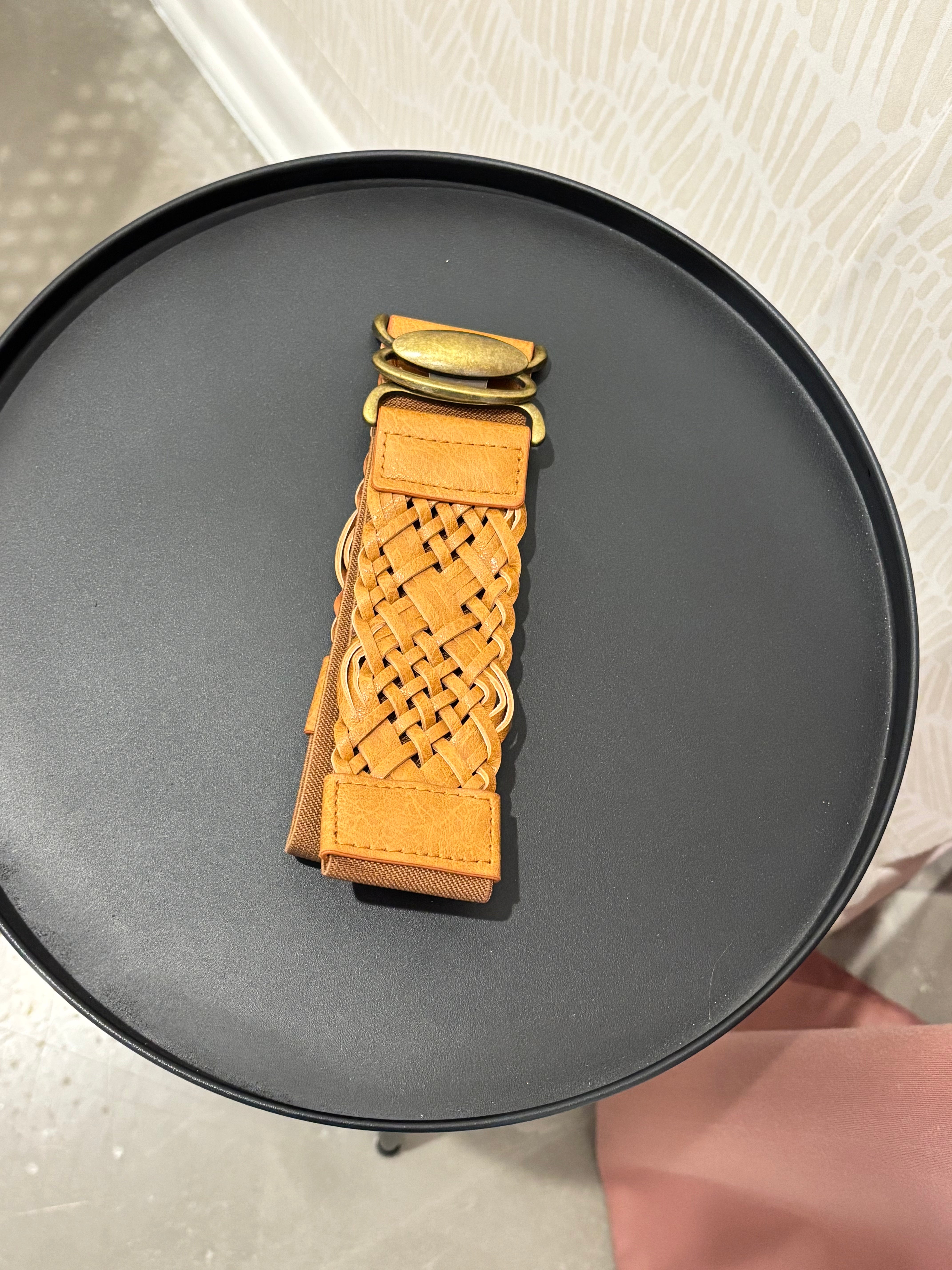 A tan woven leather belt with gold-tone hardware, displayed on a round black surface.