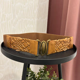 A tan leather belt with a braided woven texture and an antique bronze oval buckle, displayed on a black round side table.