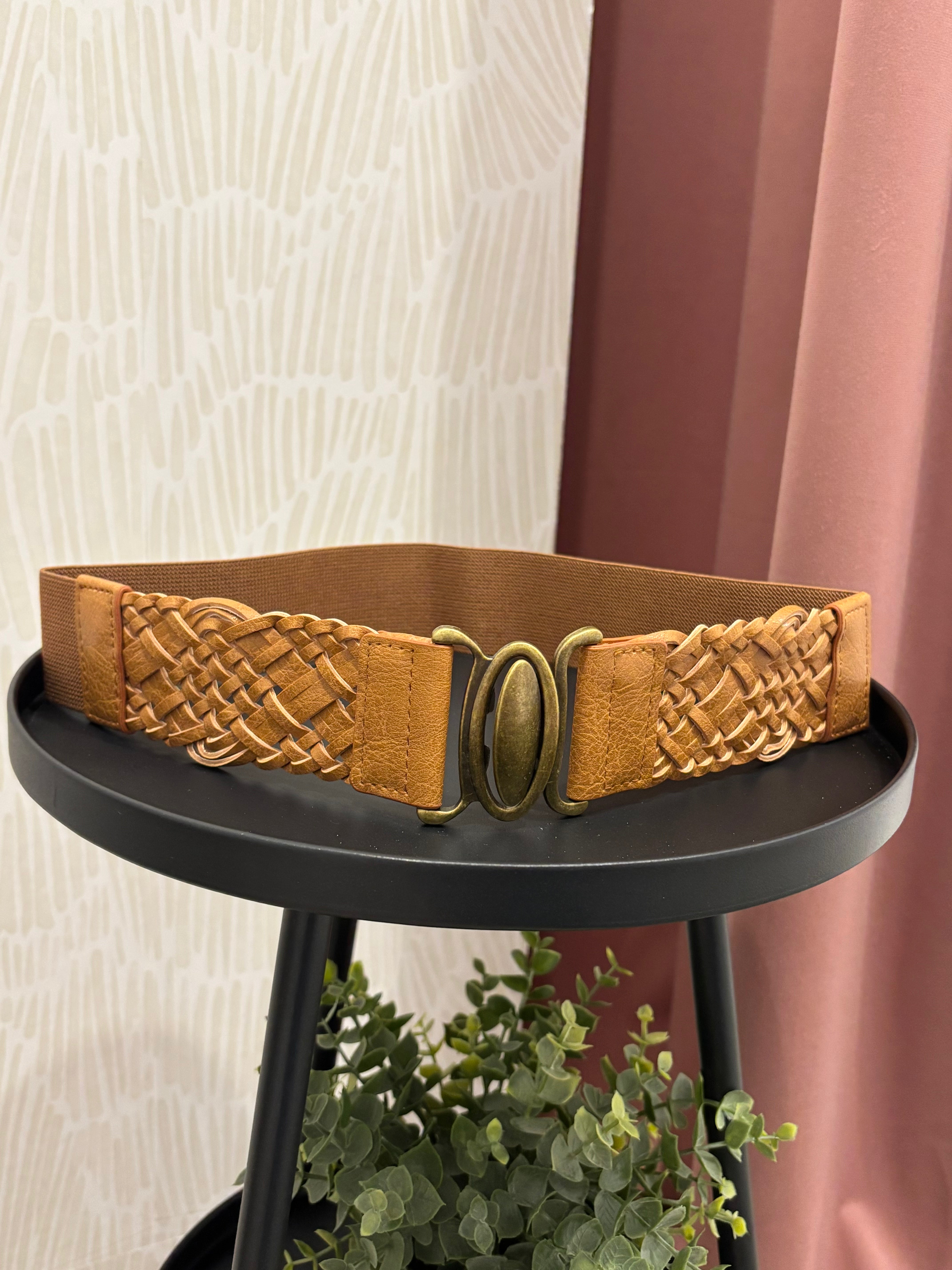 A tan leather belt with a braided woven texture and an antique bronze oval buckle, displayed on a black round side table.