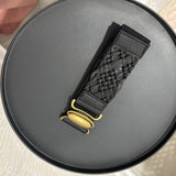 A black woven leather watch strap with a gold-tone buckle, displayed diagonally on a round black tray.
