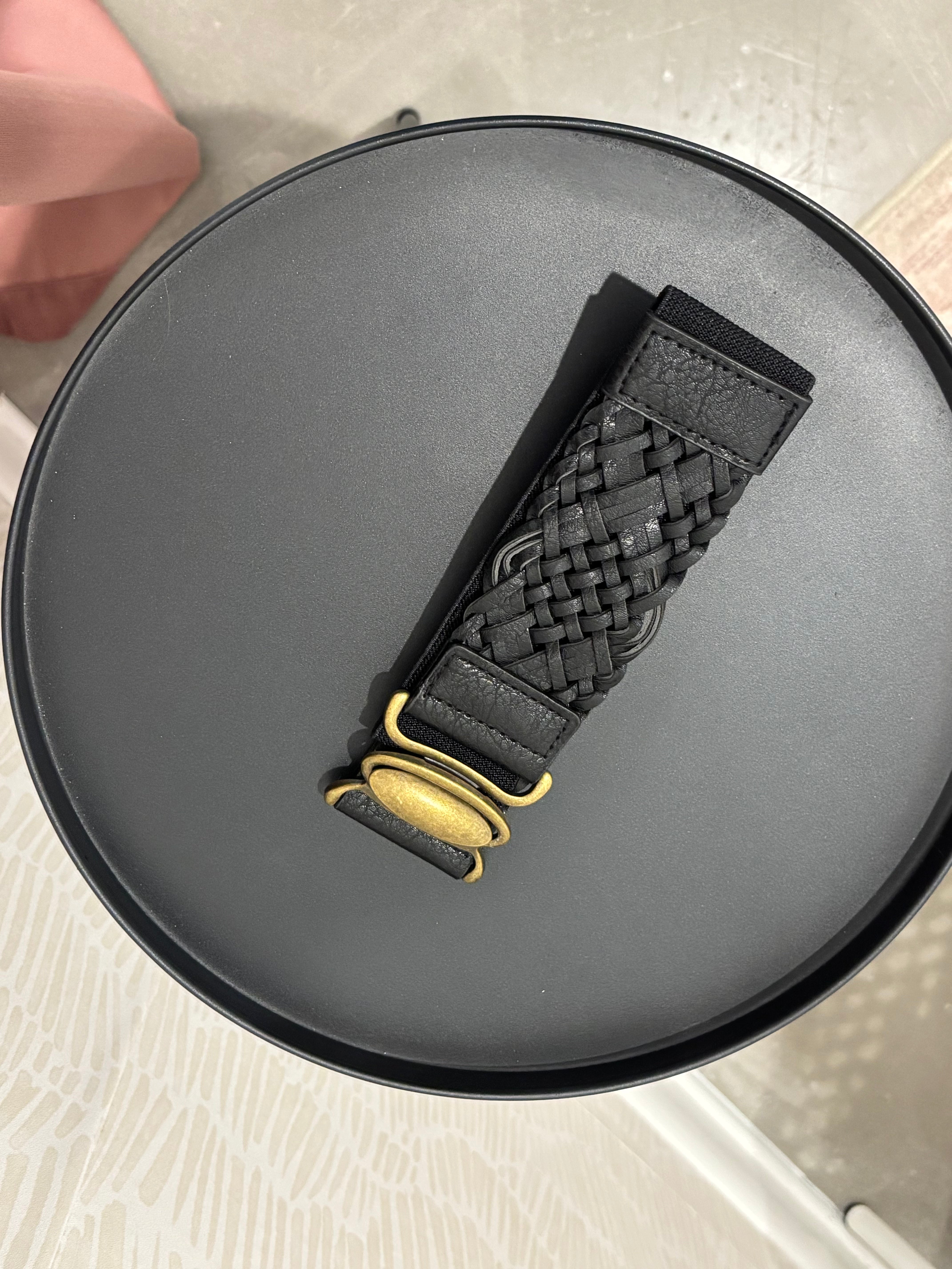 A black woven leather watch strap with a gold-tone buckle, displayed diagonally on a round black tray.