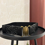 A black woven belt with a large oval gold-tone buckle, displayed on a black round table against a patterned cream wall and rust-colored curtain background.
