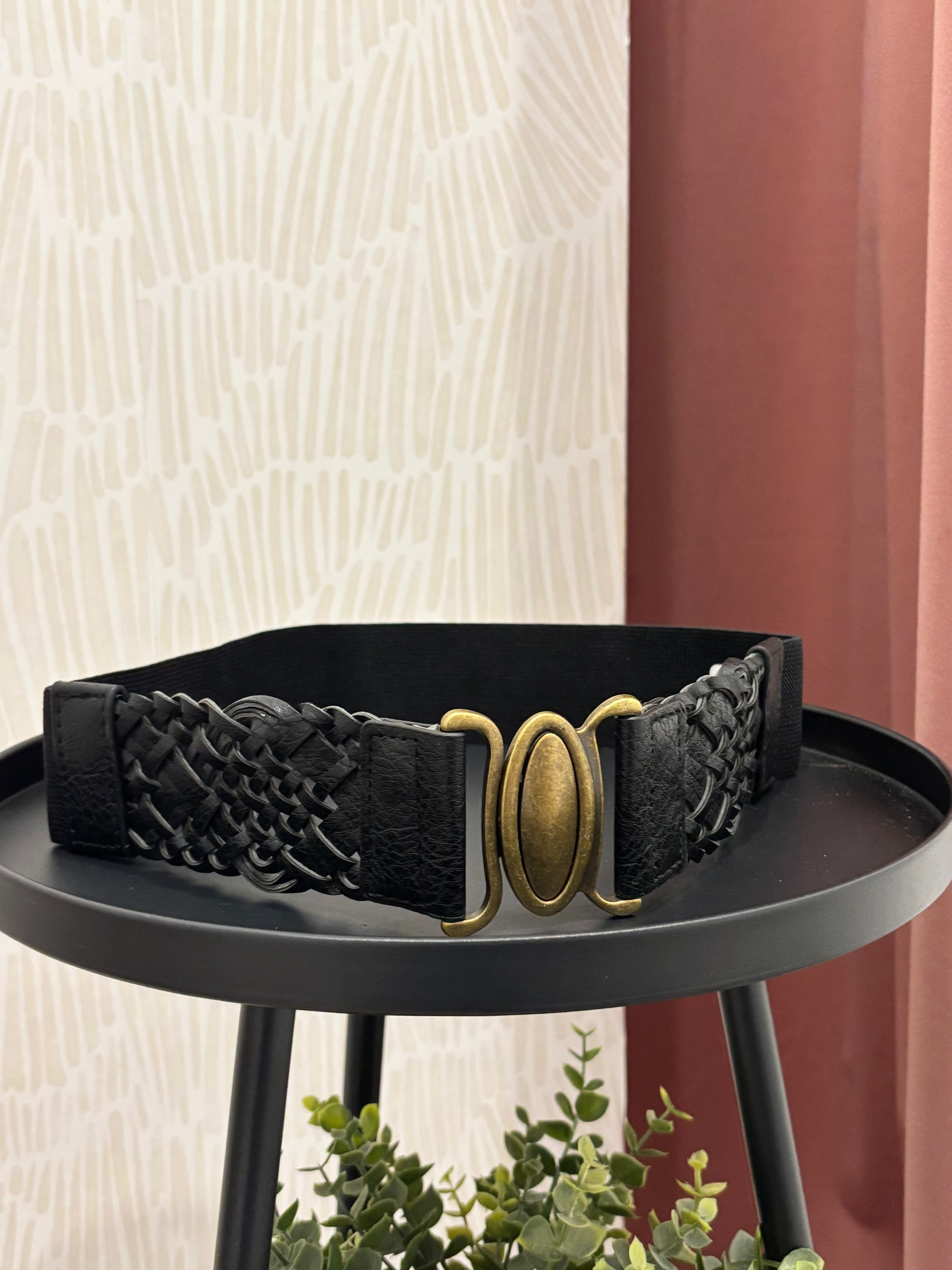 A black woven belt with a large oval gold-tone buckle, displayed on a black round table against a patterned cream wall and rust-colored curtain background.