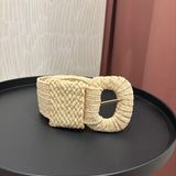 A cream-colored woven belt with a large textured oval buckle, displayed on a black round tray.