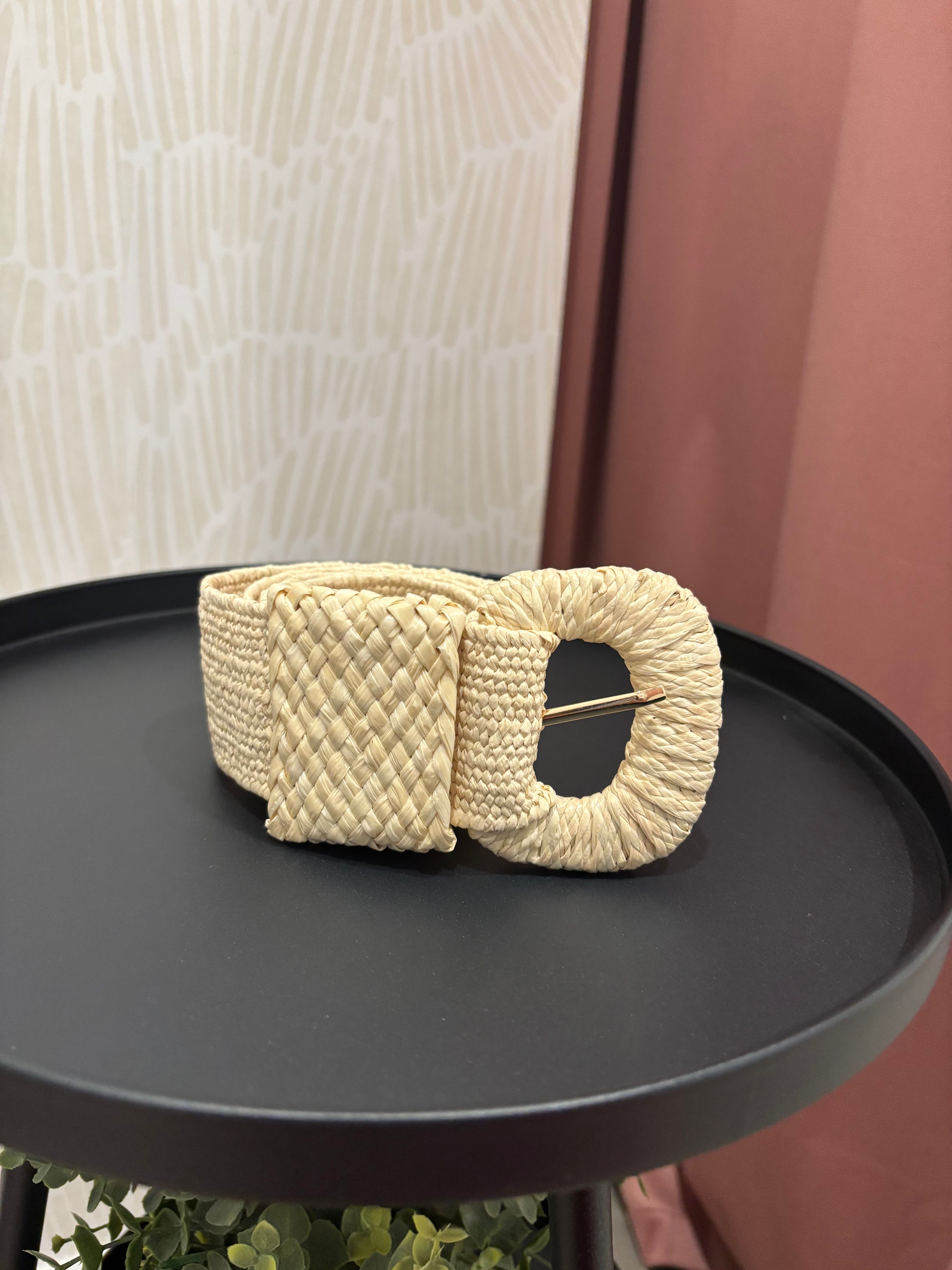 A cream-colored woven belt with a large textured oval buckle, displayed on a black round tray.