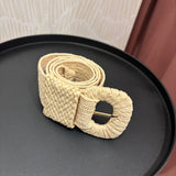A woven natural-fiber belt with a large oval buckle, resting on a round black tray. The belt has a textured, braided pattern in a light beige/cream color, with a gold-tone metal pin visible inside the buckle.
