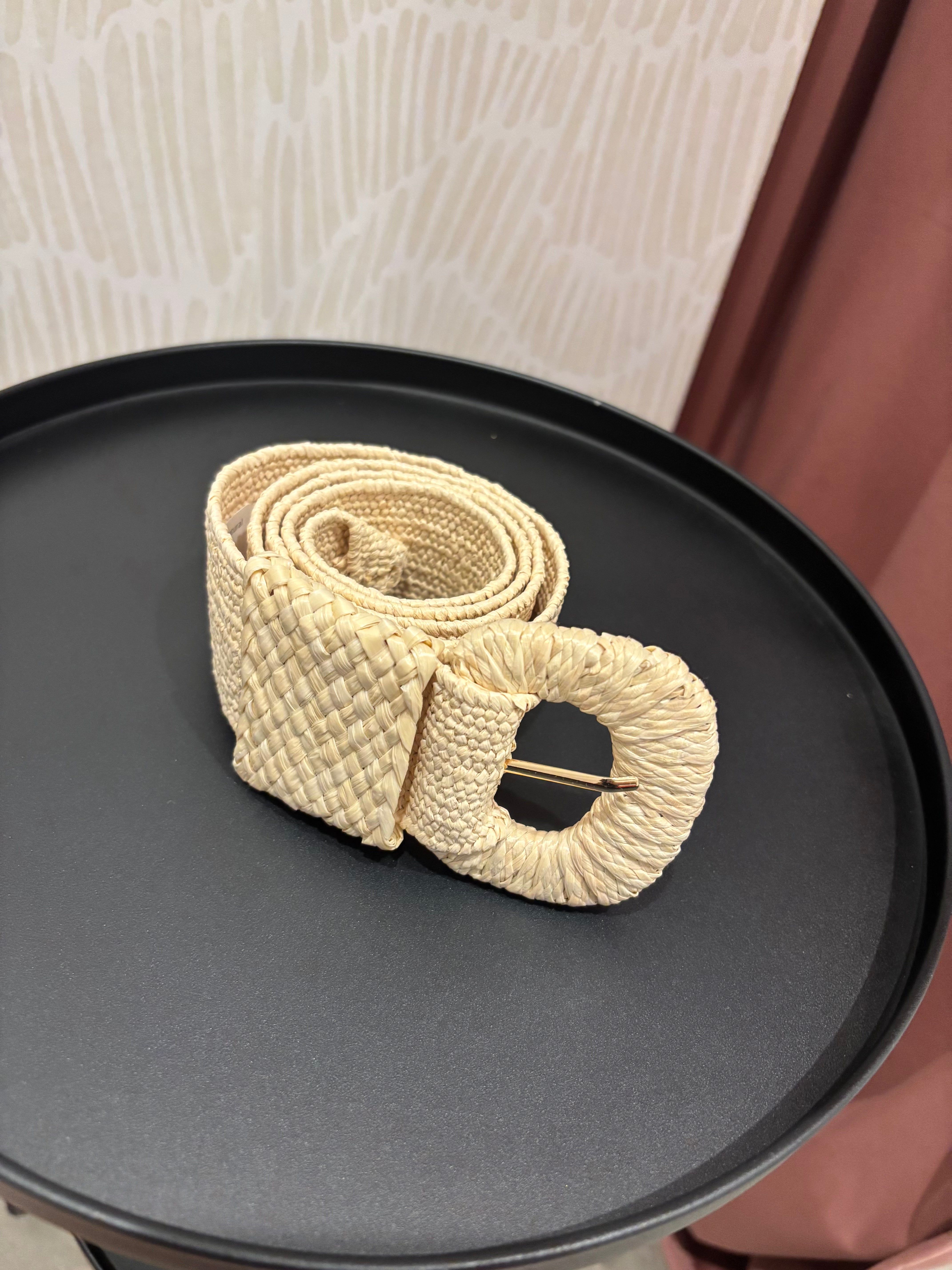 A woven natural-fiber belt with a large oval buckle, resting on a round black tray. The belt has a textured, braided pattern in a light beige/cream color, with a gold-tone metal pin visible inside the buckle.