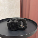 A black woven braided belt with a gold-tone metal pin buckle, displayed on a round black side table.