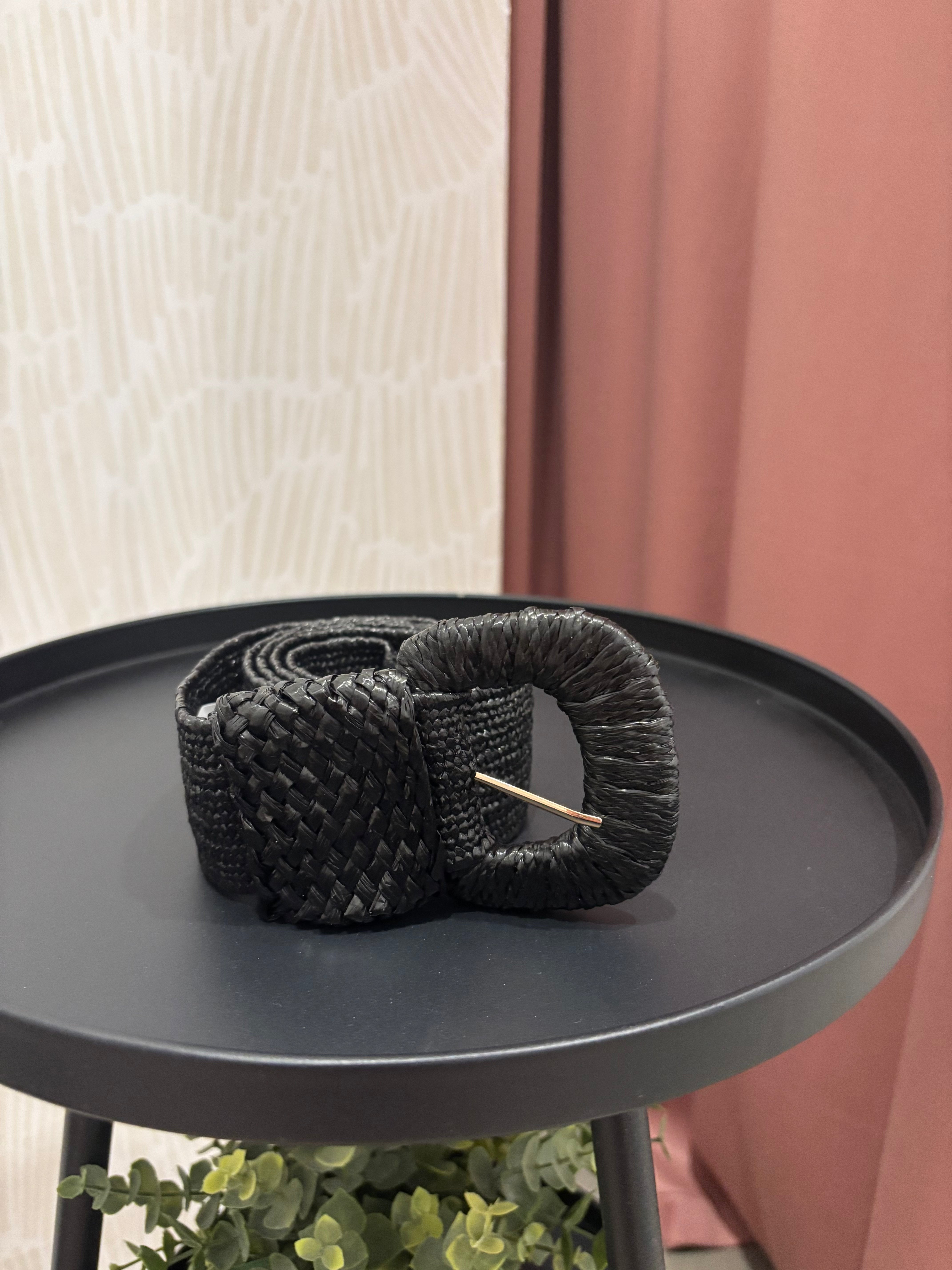 A black woven braided belt with a gold-tone metal pin buckle, displayed on a round black side table.