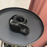 A black woven belt with a chunky oval buckle, resting on a round black side table in an indoor setting.