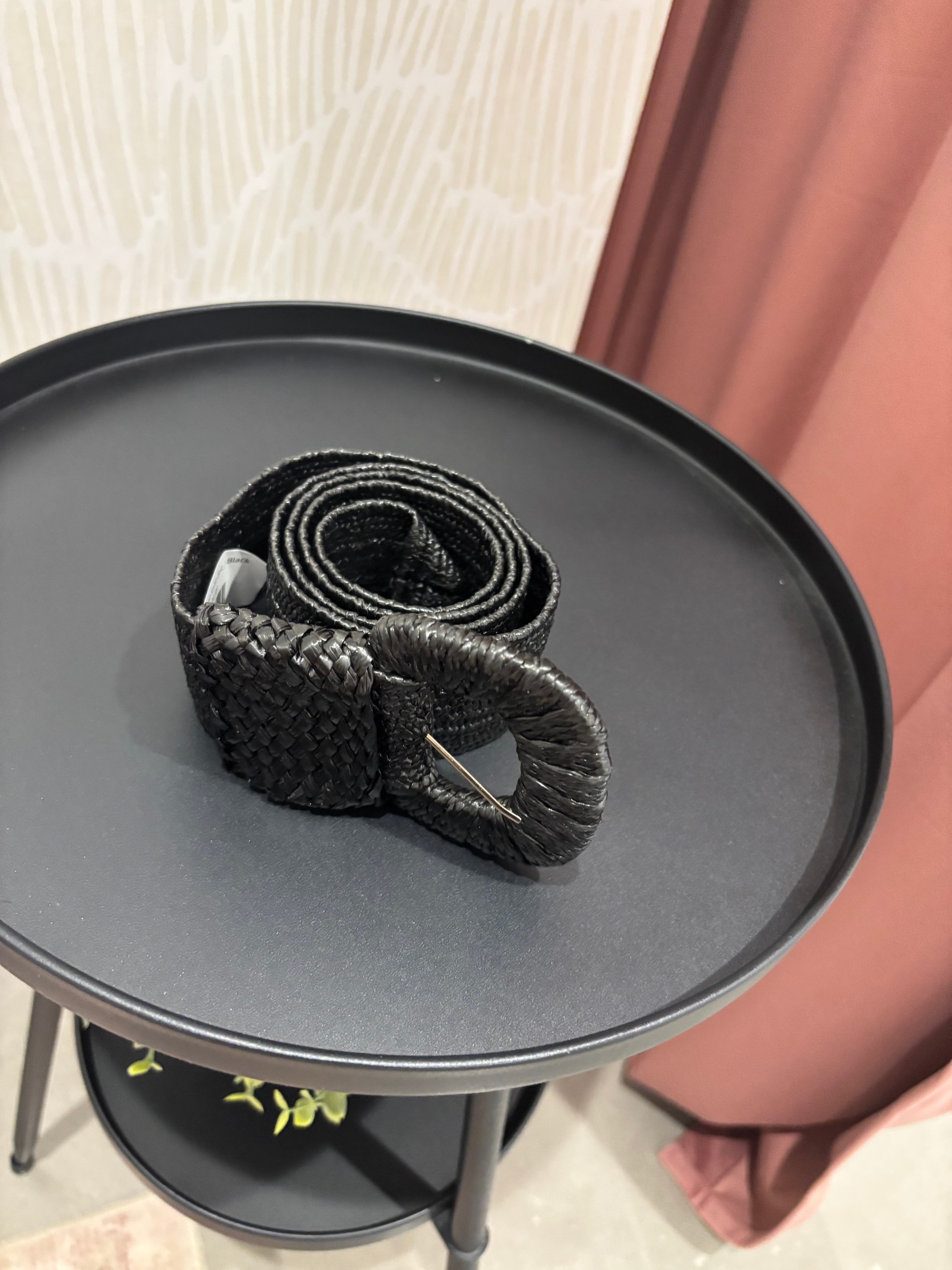A black woven belt with a chunky oval buckle, resting on a round black side table in an indoor setting.
