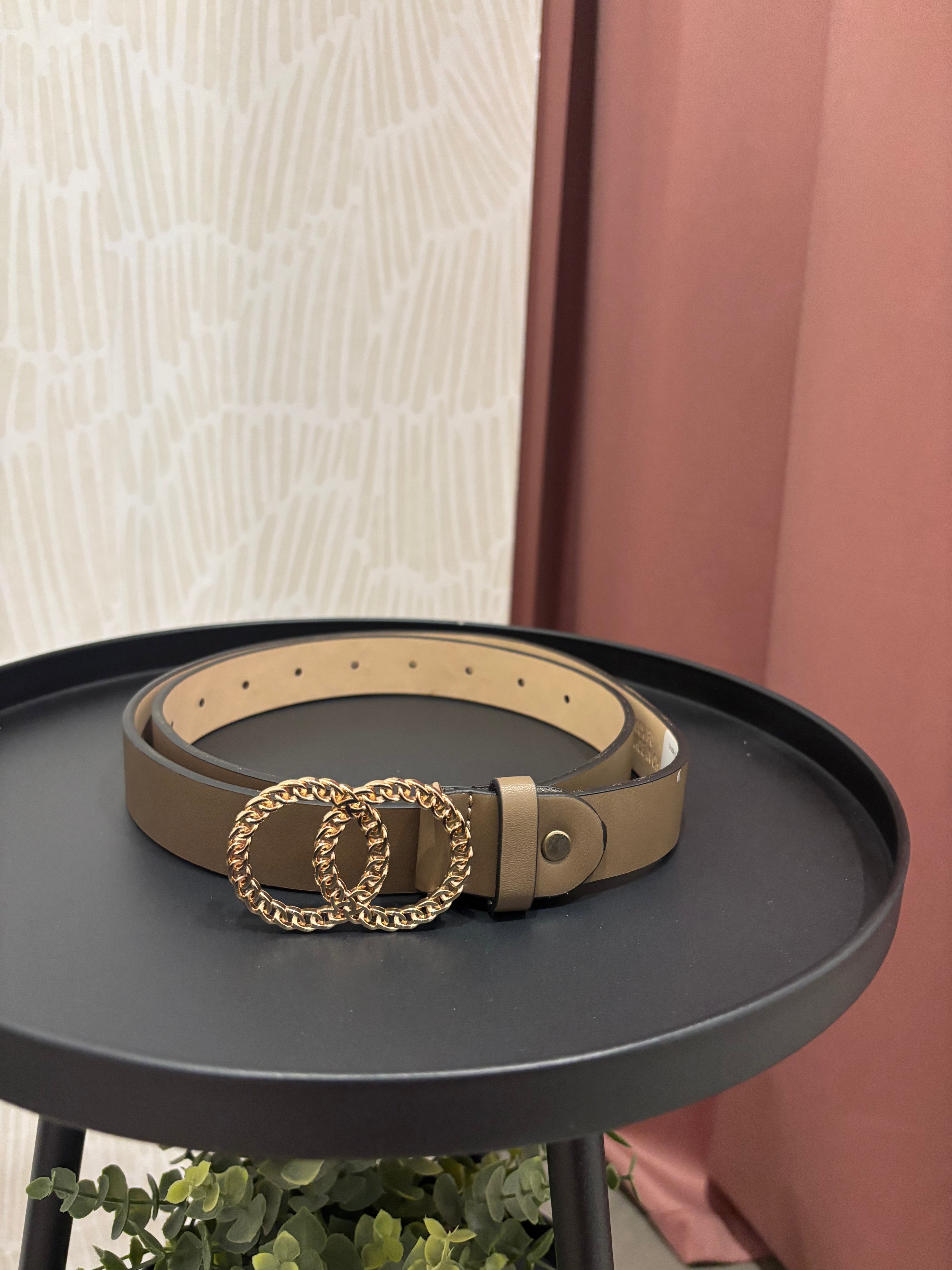 A beige leather belt with a gold-tone interlocking double-O chain buckle, displayed on a round black side table.