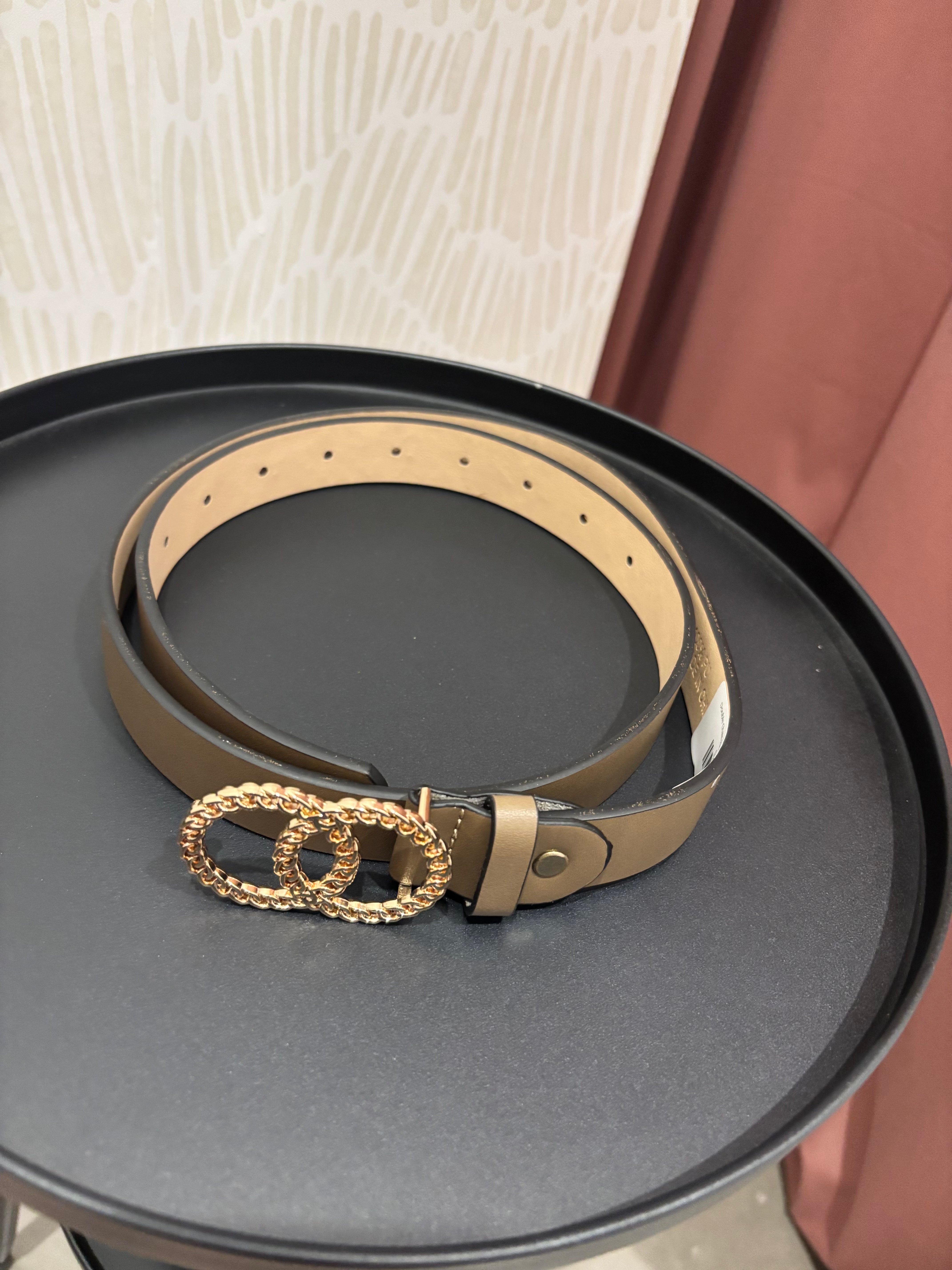 A tan leather belt with a gold-tone interlocking double-O buckle, coiled on a round black tray.