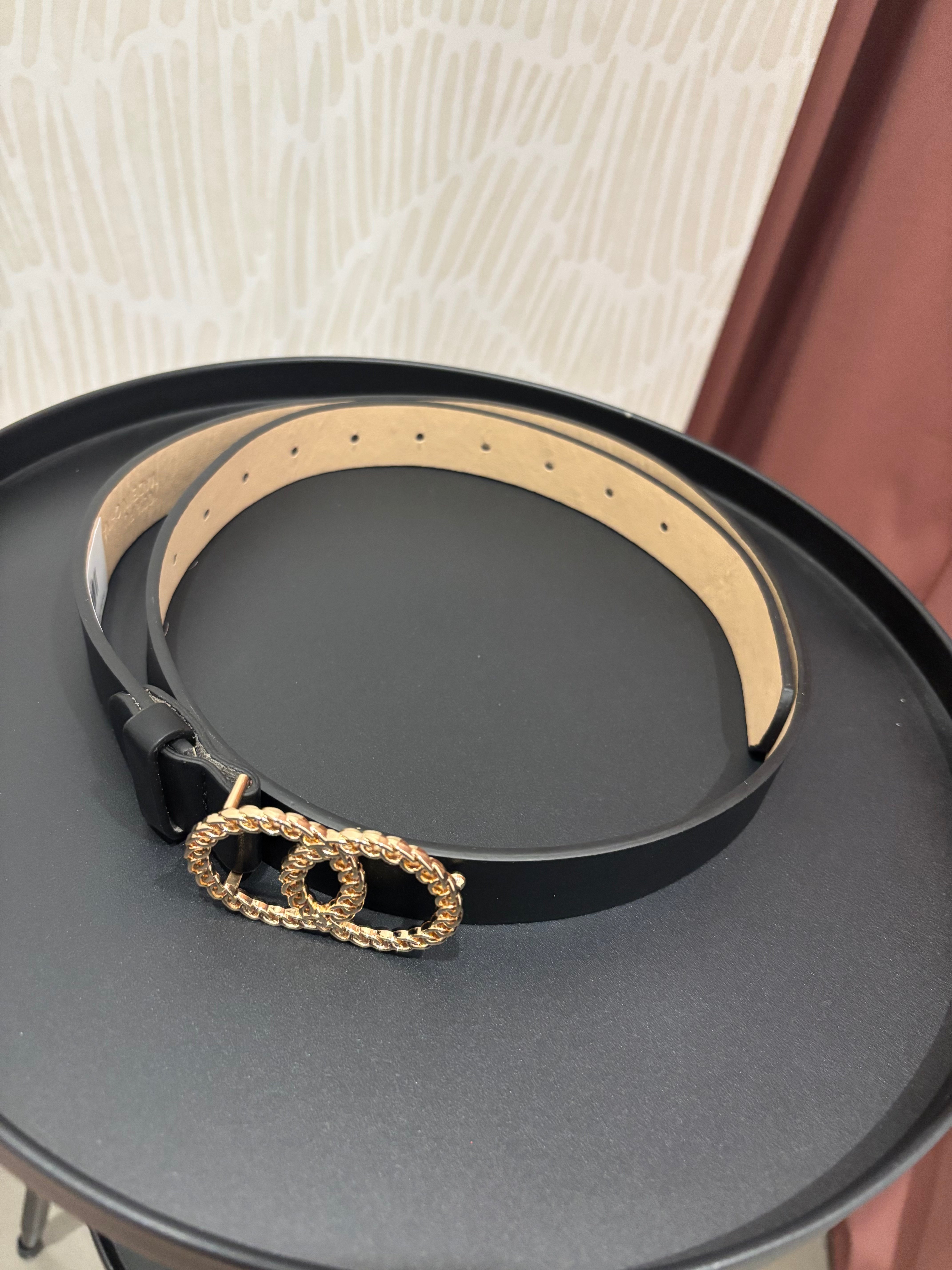 A black leather belt with a gold rope-textured double-O buckle, displayed on a round black tray.