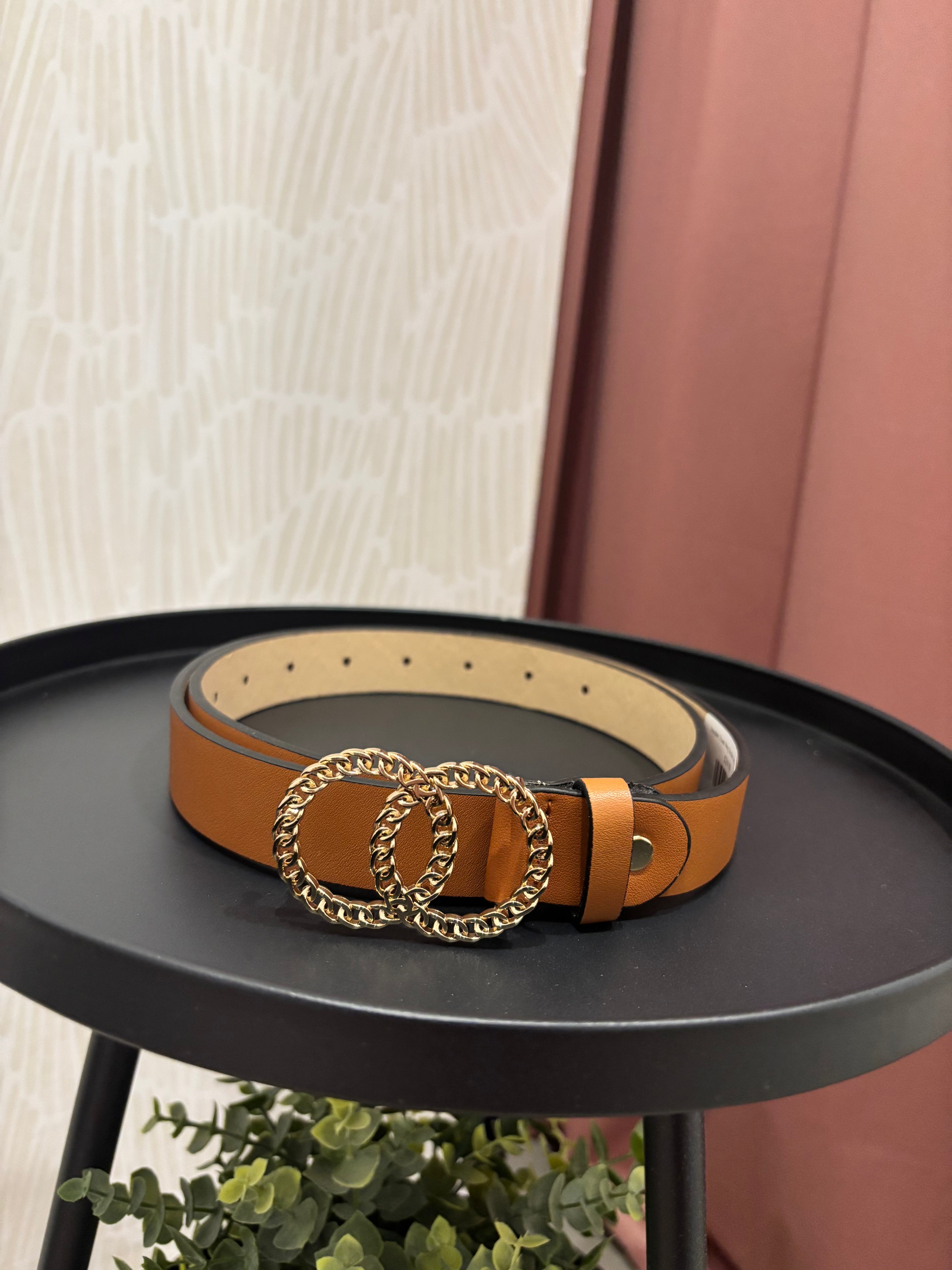 A tan leather belt with a large interlocking double-O chain buckle, displayed on a black round table.