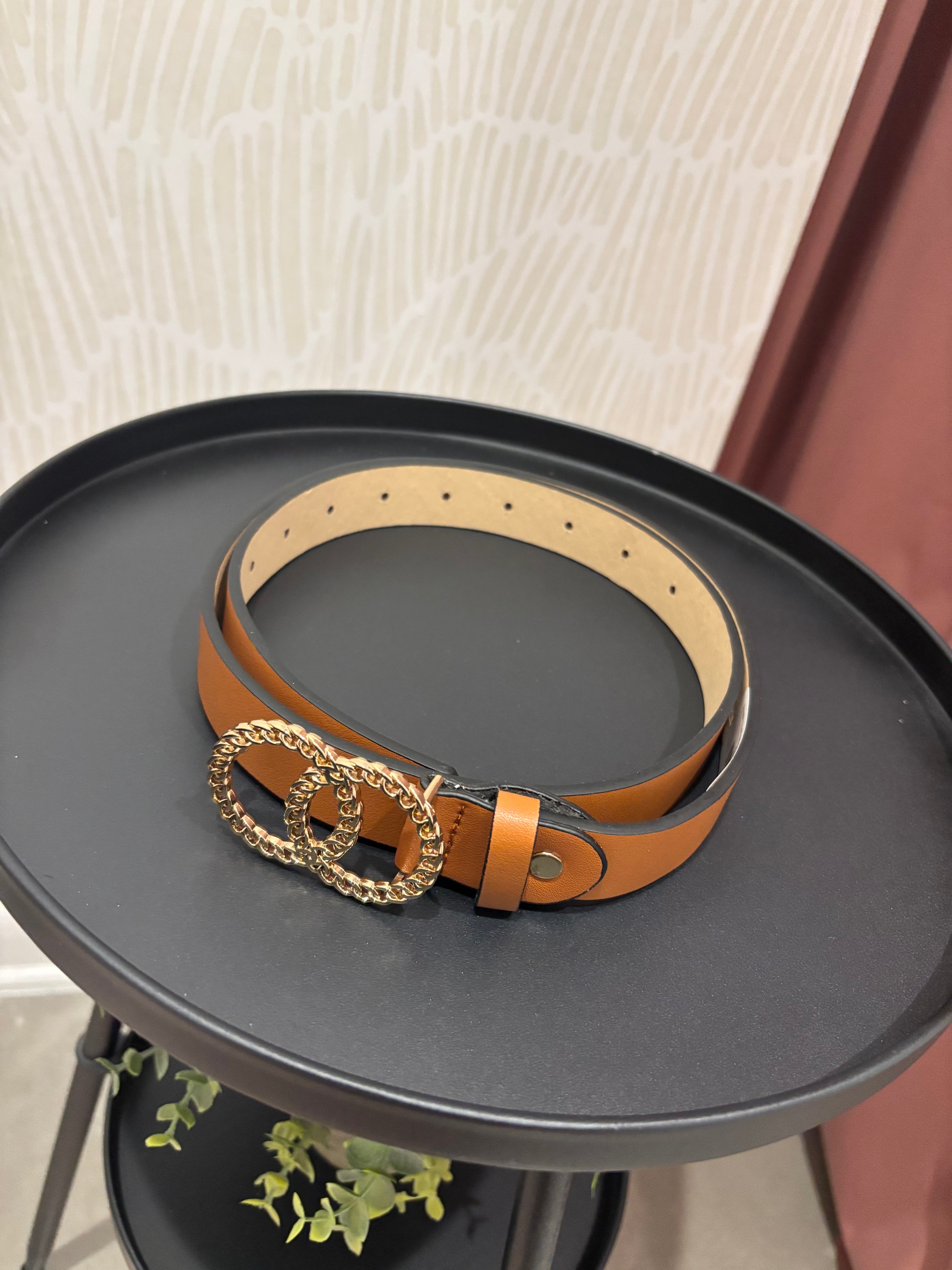 A tan leather belt with a large interlocking double-O ring buckle, displayed on a round black tray.