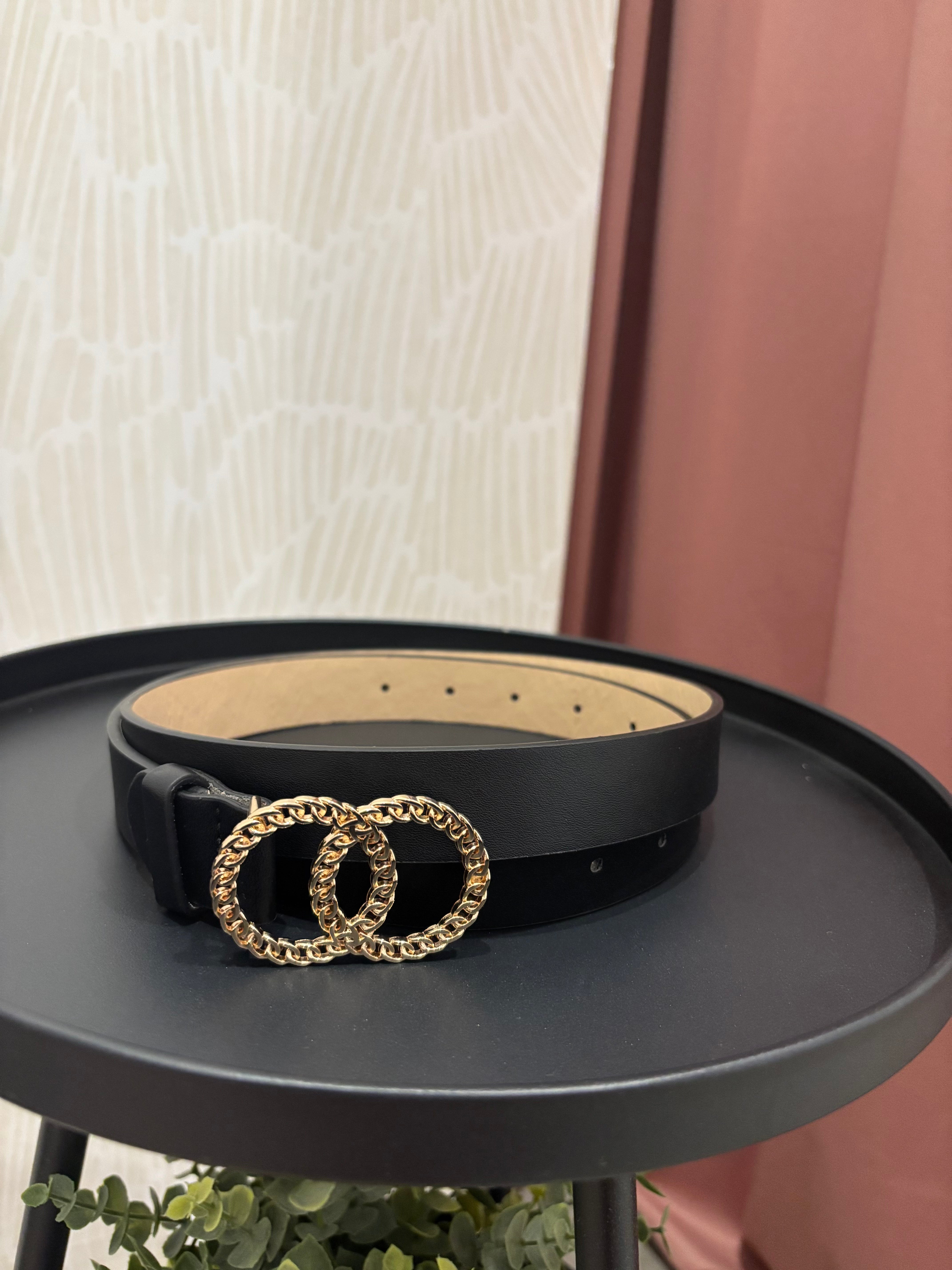 A black leather belt with a large interlocking double-ring buckle in a textured gold-tone finish, displayed on a round black side table.