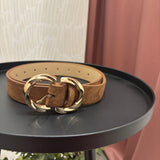 A tan suede belt with a large, polished gold-tone double-ring buckle, displayed on a round black side table.