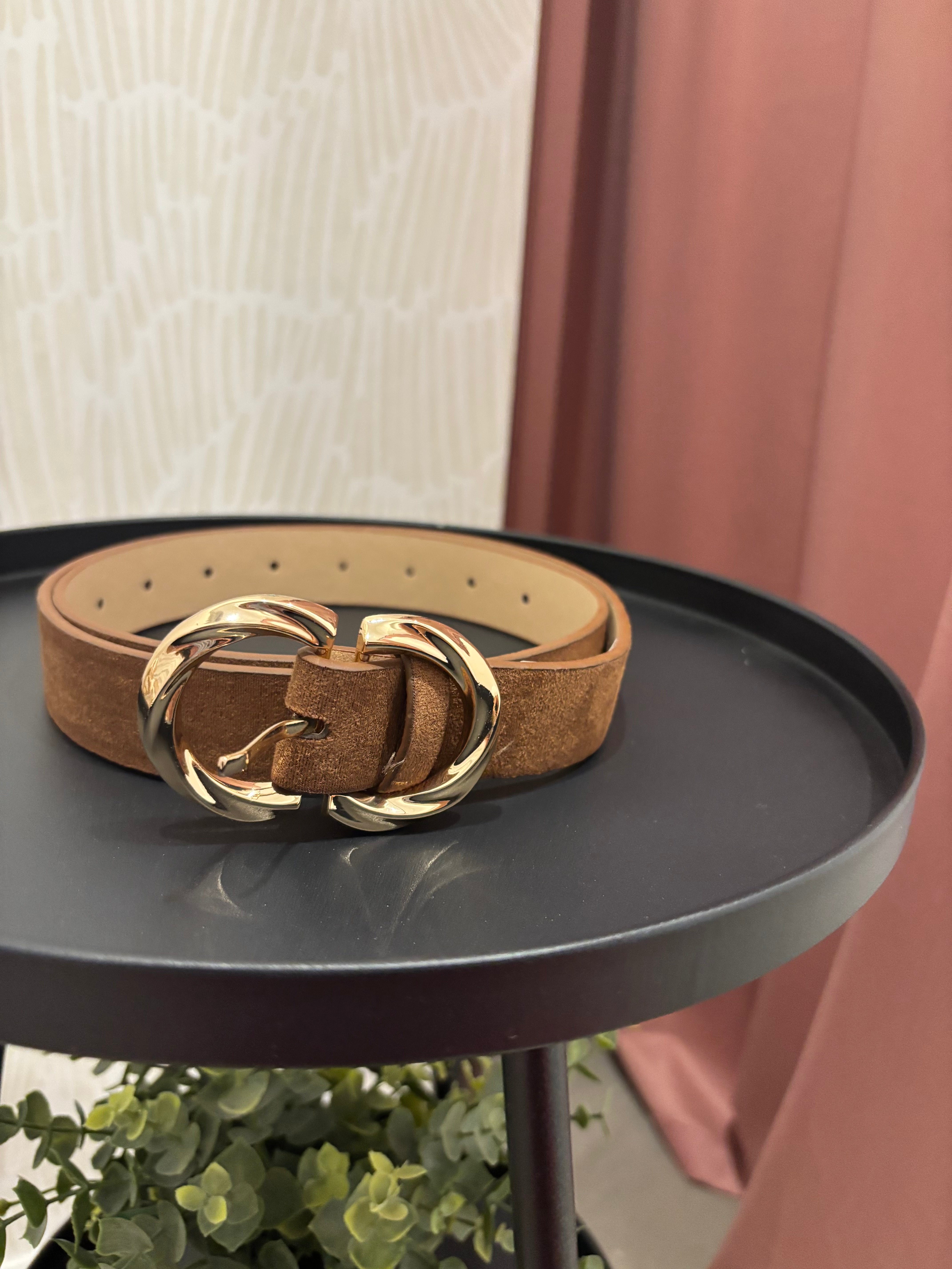 A tan suede belt with a large, polished gold-tone double-ring buckle, displayed on a round black side table.