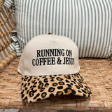 A baseball cap resting on a woven rattan chair in front of a striped pillow. The cap has an off-white crown with black embroidered text reading 'RUNNING ON COFFEE & JESUS' and a tan leopard-print brim.