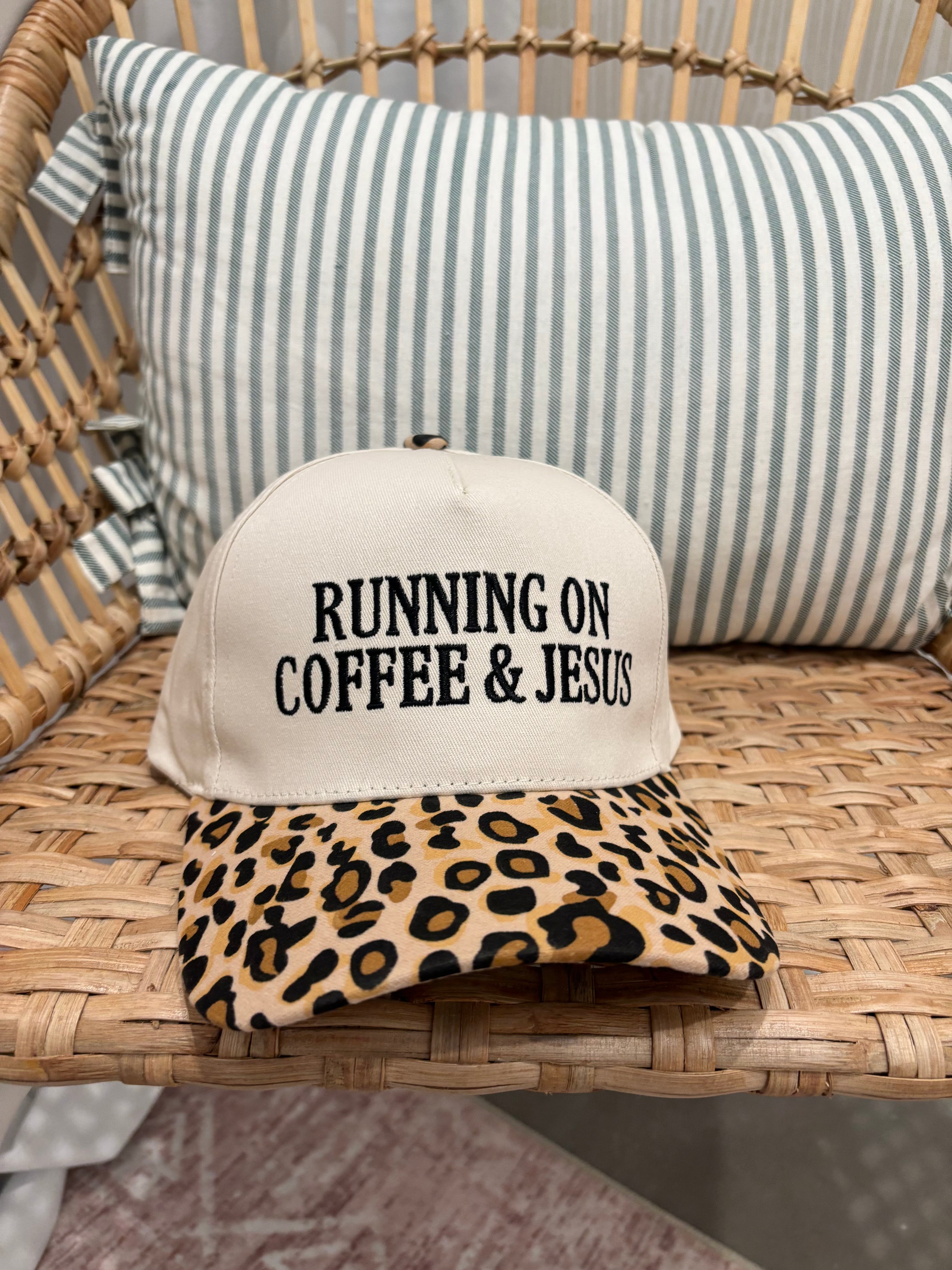 A baseball cap resting on a woven rattan chair in front of a striped pillow. The cap has an off-white crown with black embroidered text reading 'RUNNING ON COFFEE & JESUS' and a tan leopard-print brim.