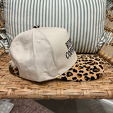 A baseball cap resting on a woven rattan chair, featuring a cream-colored crown with black embroidered text and a leopard-print visor.