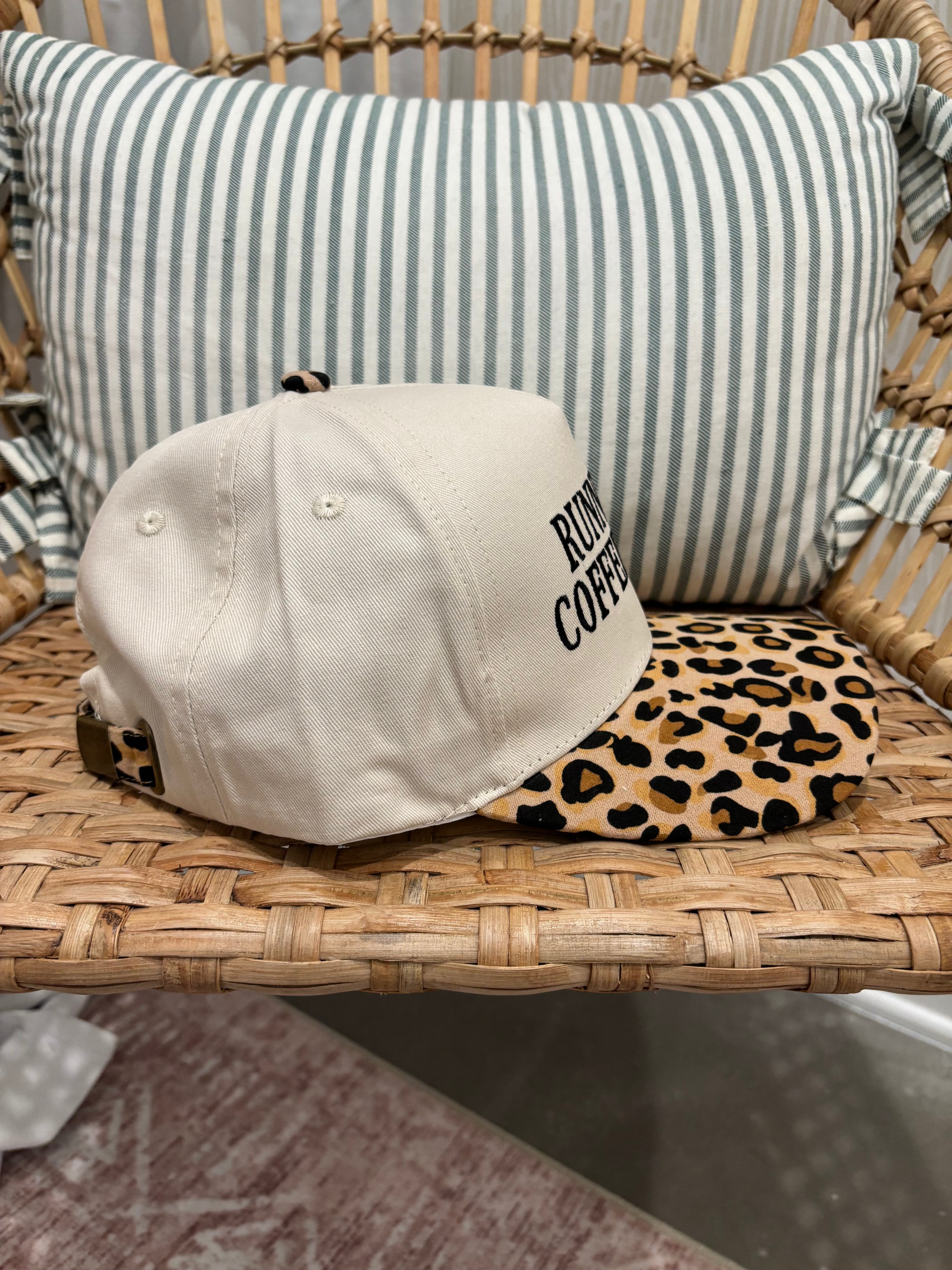 A baseball cap resting on a woven rattan chair, featuring a cream-colored crown with black embroidered text and a leopard-print visor.