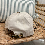 A beige baseball cap with a leopard-print visor and matching leopard-print back strap, resting on a woven rattan chair next to a striped pillow.