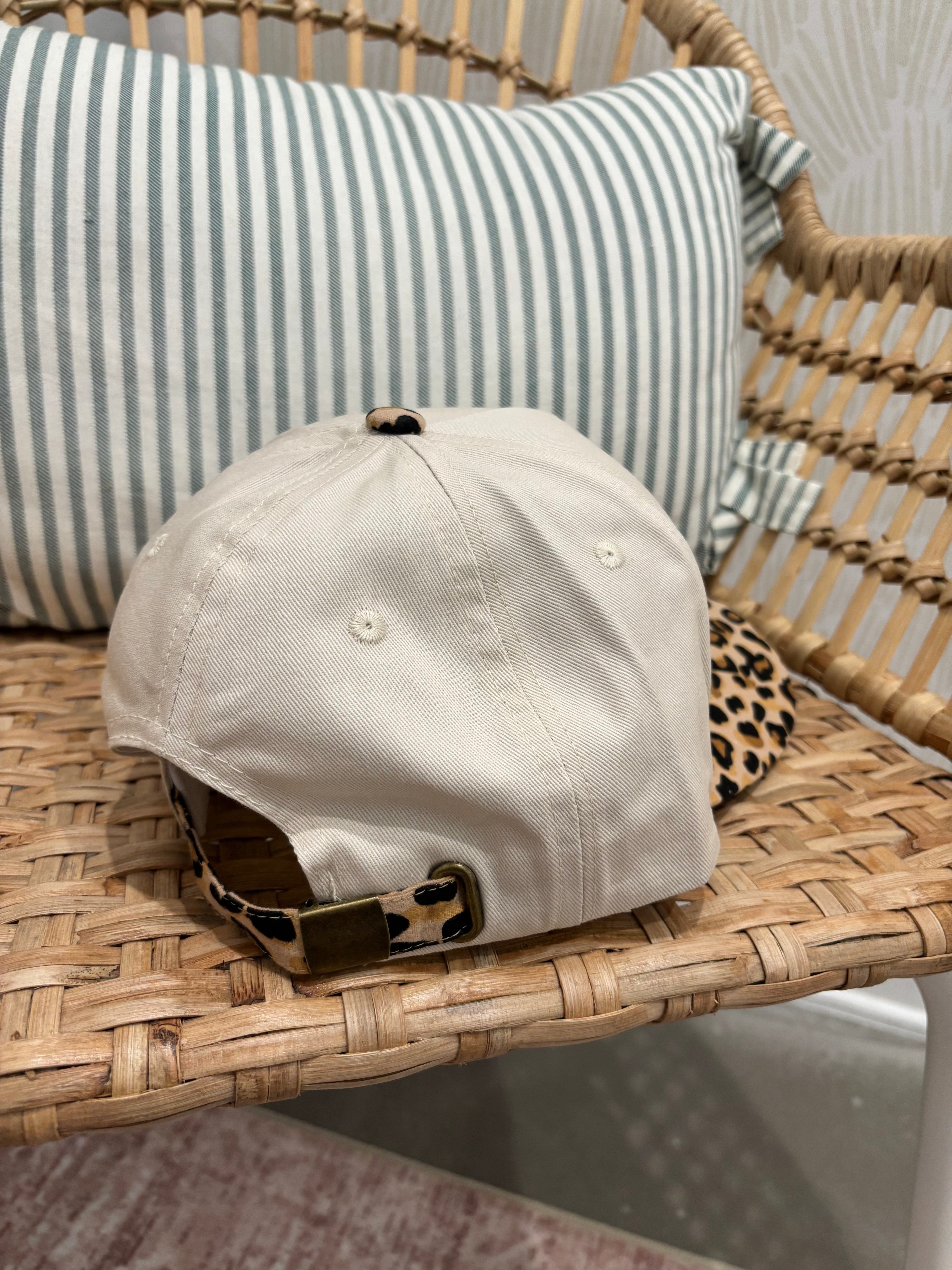 A beige baseball cap with a leopard-print visor and matching leopard-print back strap, resting on a woven rattan chair next to a striped pillow.