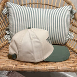 A beige baseball cap with a dark green visor and green strap detail, resting on a woven rattan chair in front of a green and white striped pillow.