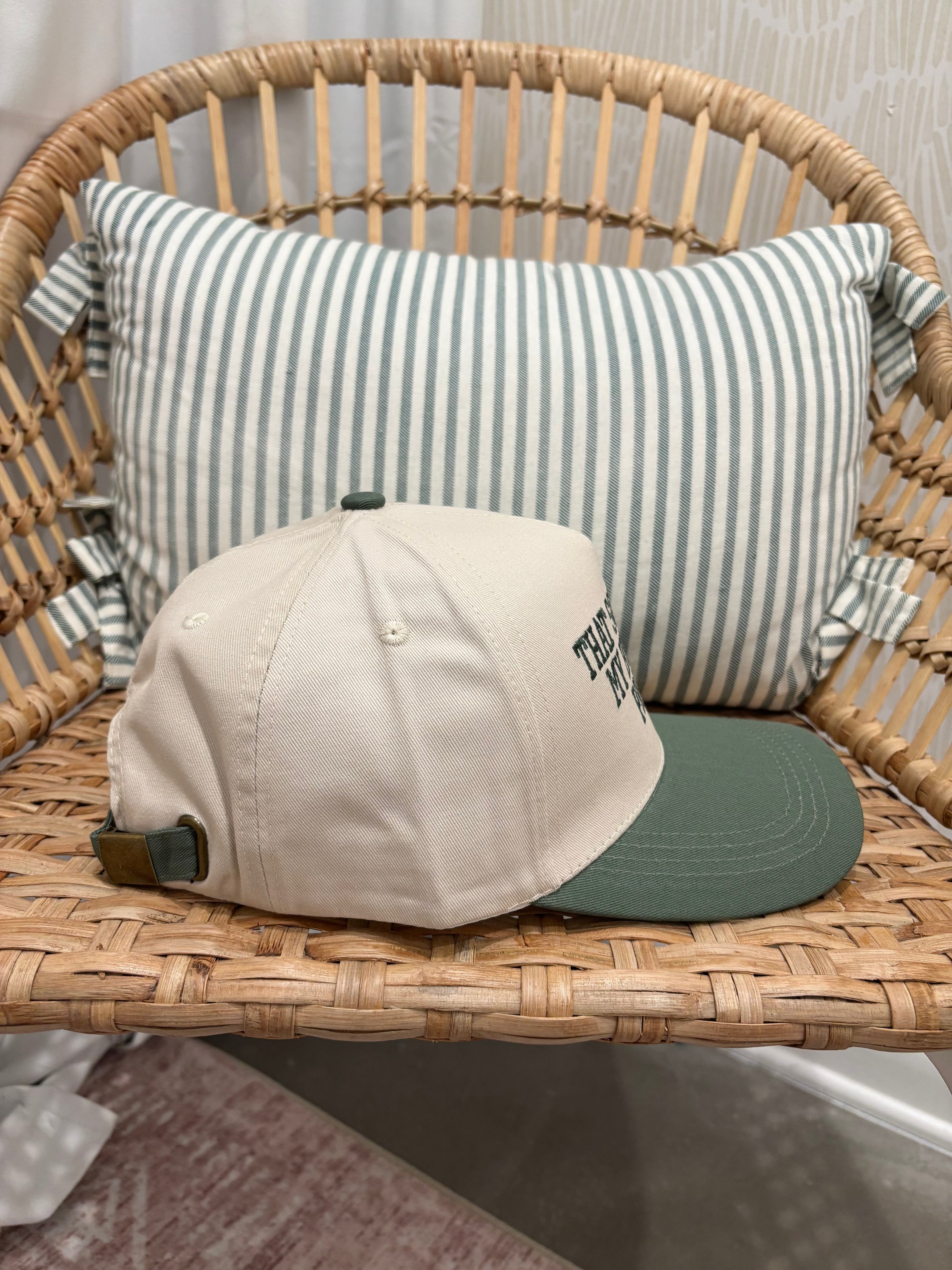 A beige baseball cap with a dark green visor and green strap detail, resting on a woven rattan chair in front of a green and white striped pillow.