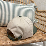 A beige baseball cap with a dark green visor and green strap detail, resting on a woven rattan chair in front of a green and white striped pillow.