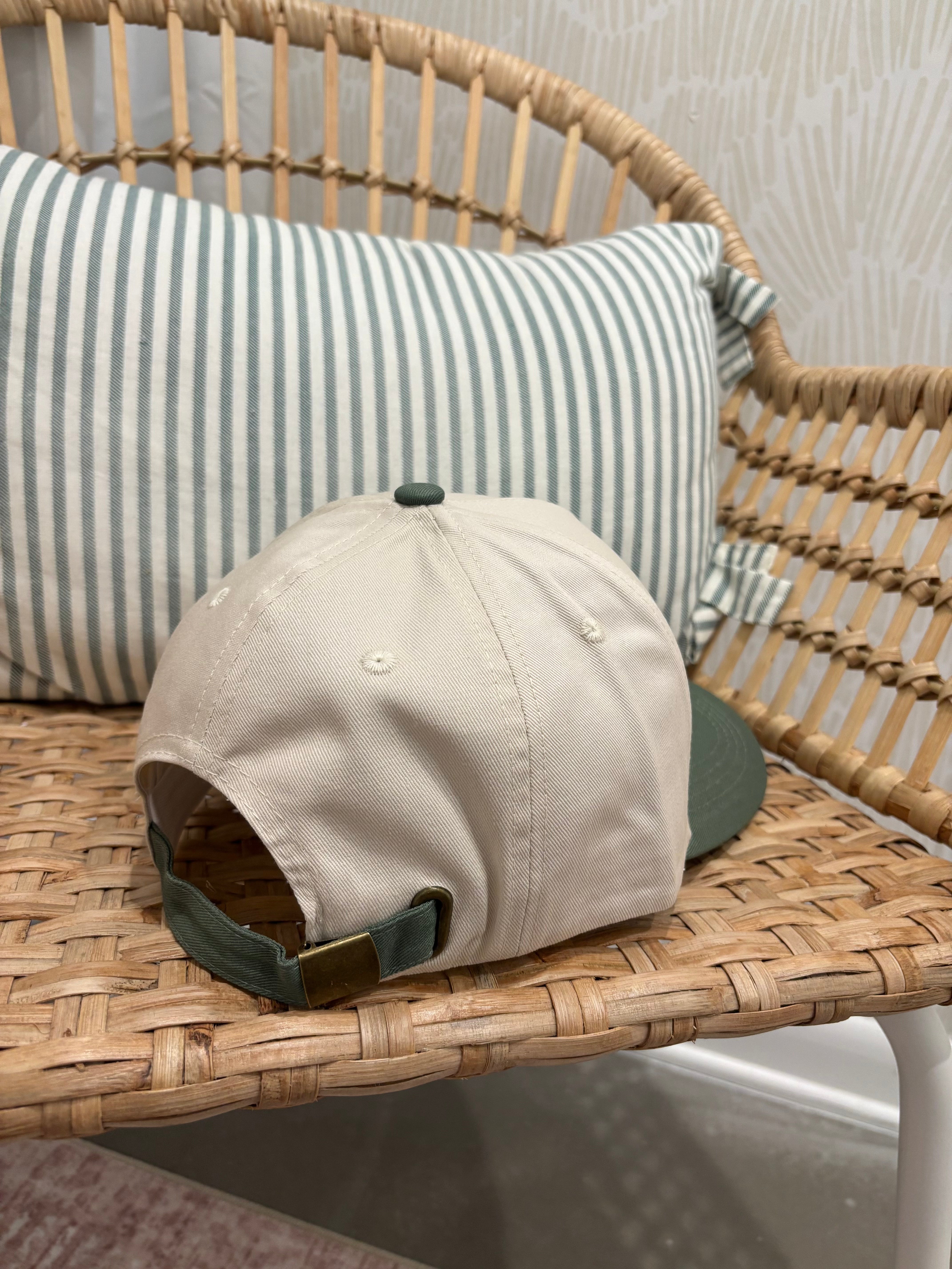 A beige baseball cap with a dark green visor and green strap detail, resting on a woven rattan chair in front of a green and white striped pillow.