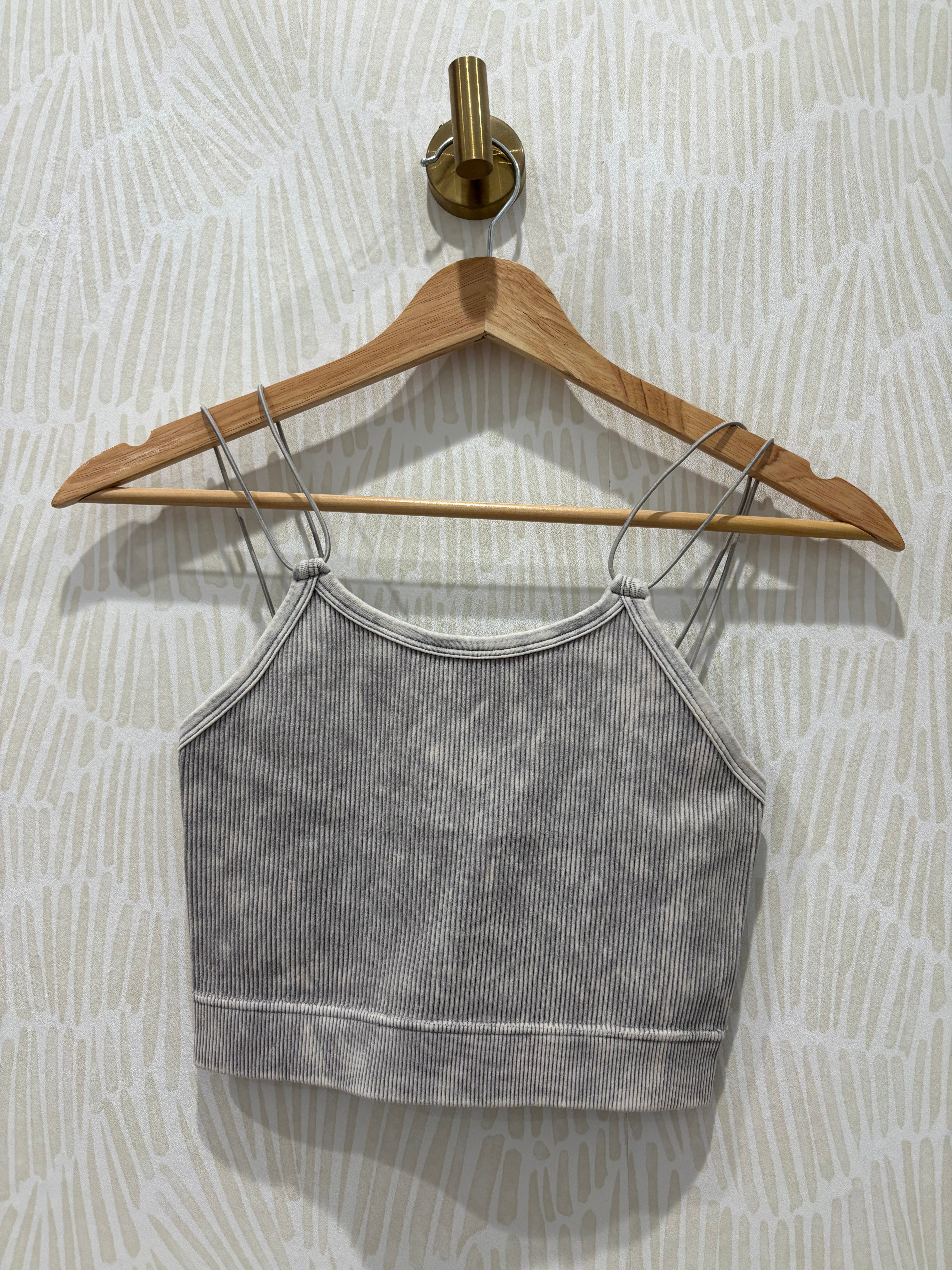 A gray ribbed knit camisole crop top with thin shoulder straps, hanging on a wooden hanger against a light patterned wallpaper background.