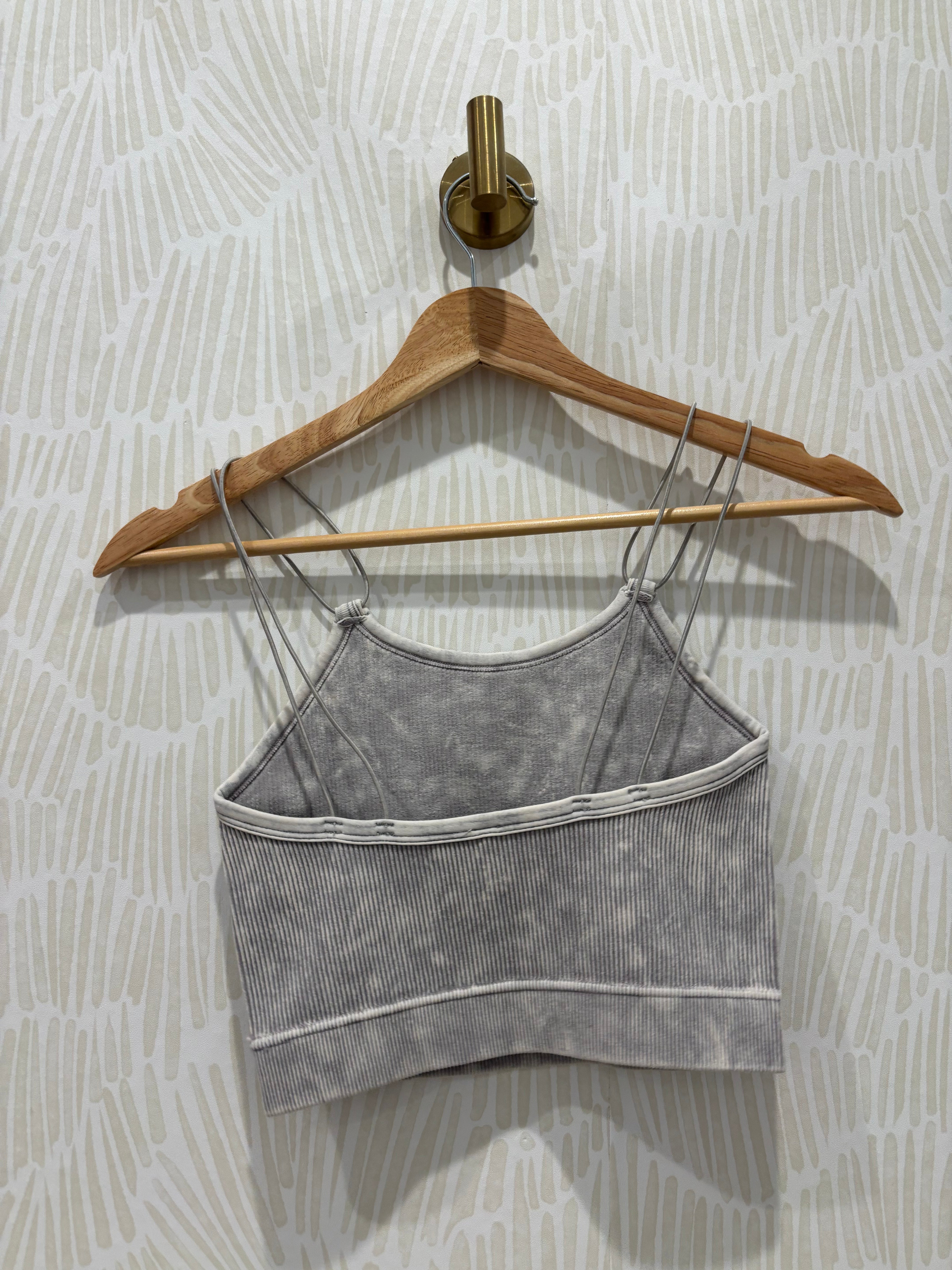 A gray ribbed knit camisole tank top with thin straps and horizontal seam detailing, hanging on a wooden hanger against a patterned wallpaper background.
